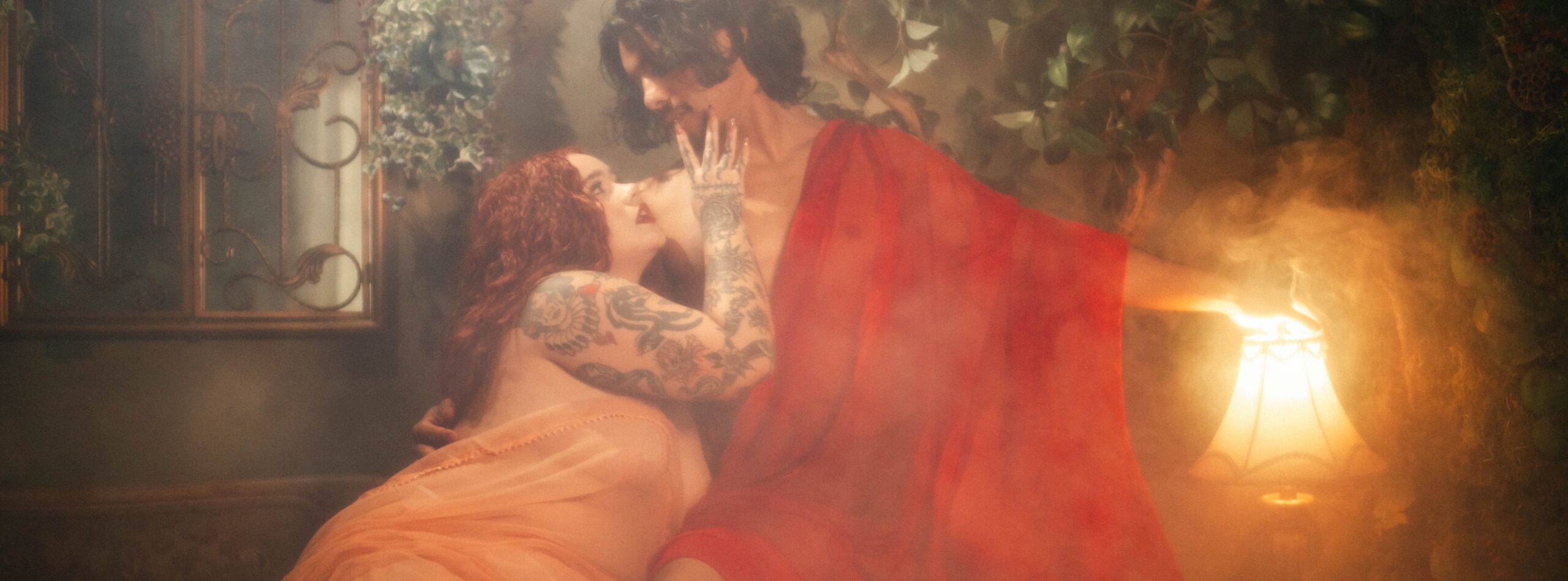 A couple embraces during a Renaissance boudoir photoshoot at Royal Lune Studio, the woman draped in wet coral chiffon while the man wraps himself in wet red silk and reaches toward a glowing fringed lamp, surrounded by fog, ivy, and a vintage wrought iron mirror, Royal Lune Photo.