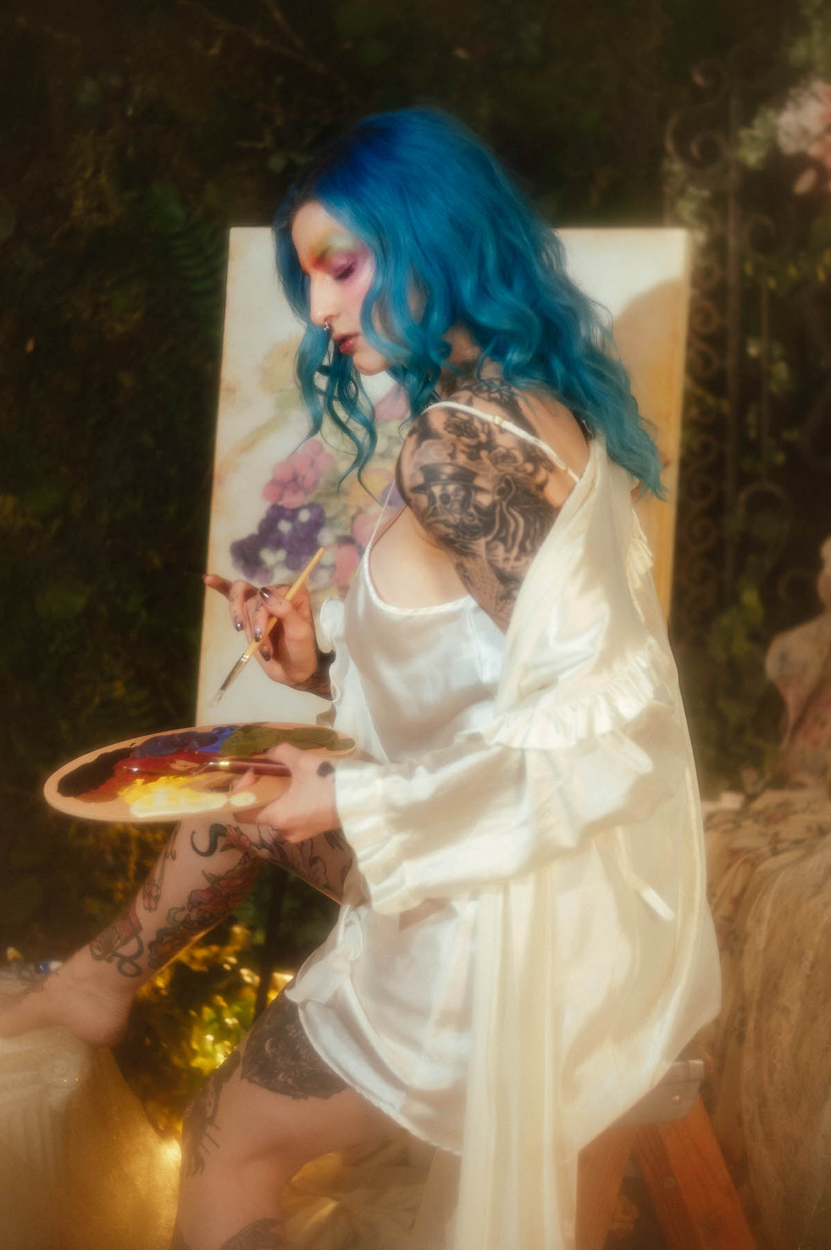 A woman in an ivory vintage slip dress paints a floral still life canvas on an easel while holding a loaded palette, photographed by a Dallas fine art photographer at Royal Lune Photo.