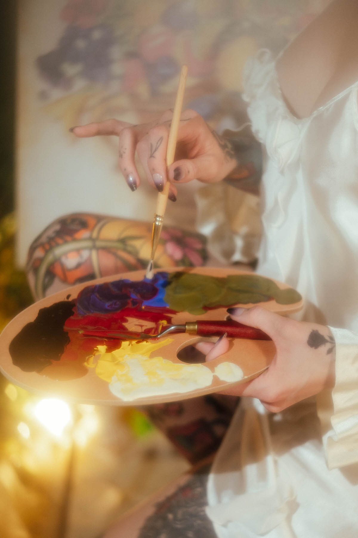 A tattooed hand holds a wooden painter's palette loaded with blue, purple, red, yellow, and black paint while another hand raises a paintbrush during this artist boudoir photoshoot at Royal Lune Photo.