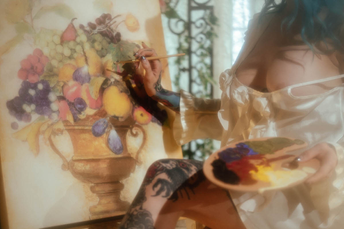 A woman paints a fruit and urn still life canvas on an easel while holding a wooden palette, her tattooed arm visible beneath an ivory ruffled sleeve, photographed by a Dallas fine art photographer at Royal Lune Photo.