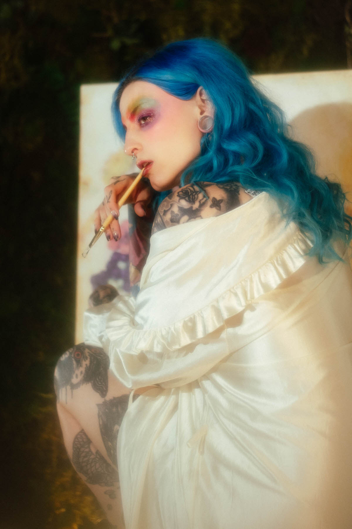 Profile portrait of a woman with blue hair holding a paintbrush near her lips, wearing an ivory ruffled robe with warm golden light behind her, taken during a creative portrait photography session at Royal Lune Photo.