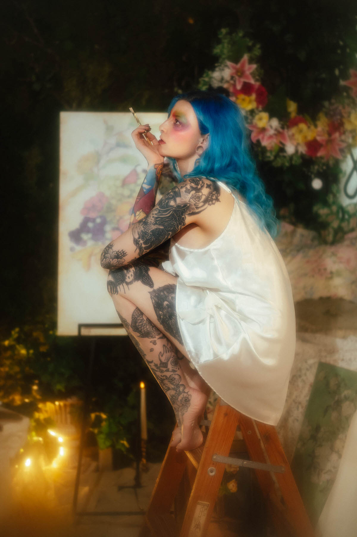 A woman with blue hair sits on a vintage wooden step ladder in an ivory slip dress, paintbrush held near her chin, with candlelight and florals glowing behind her during this artist boudoir photoshoot at Royal Lune Photo.