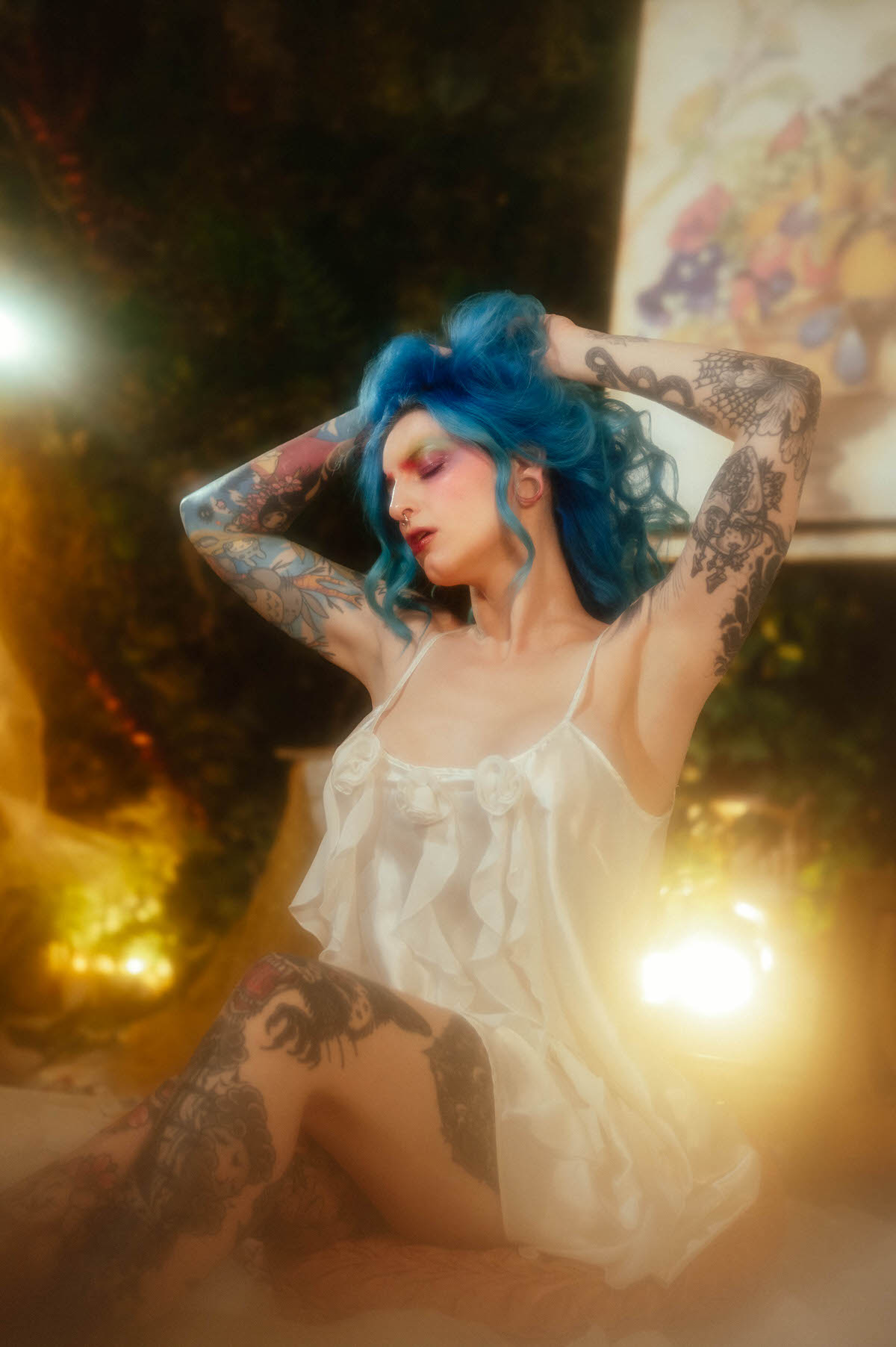 A woman kneeling in an ivory slip dress raises both arms to hold up her blue hair, eyes closed, with warm golden light and a floral still life painting behind her during this artist boudoir photoshoot at Royal Lune Photo.