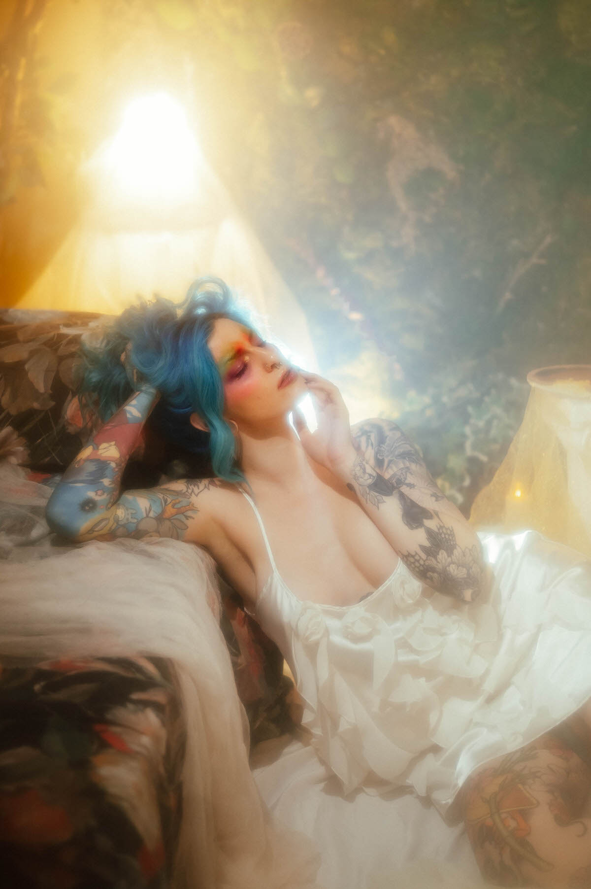 A woman with blue hair reclines against a floral backdrop with her head tilted back and eyes closed, wearing an ivory slip dress lit by warm golden backlight during this artist boudoir photoshoot at Royal Lune Photo.