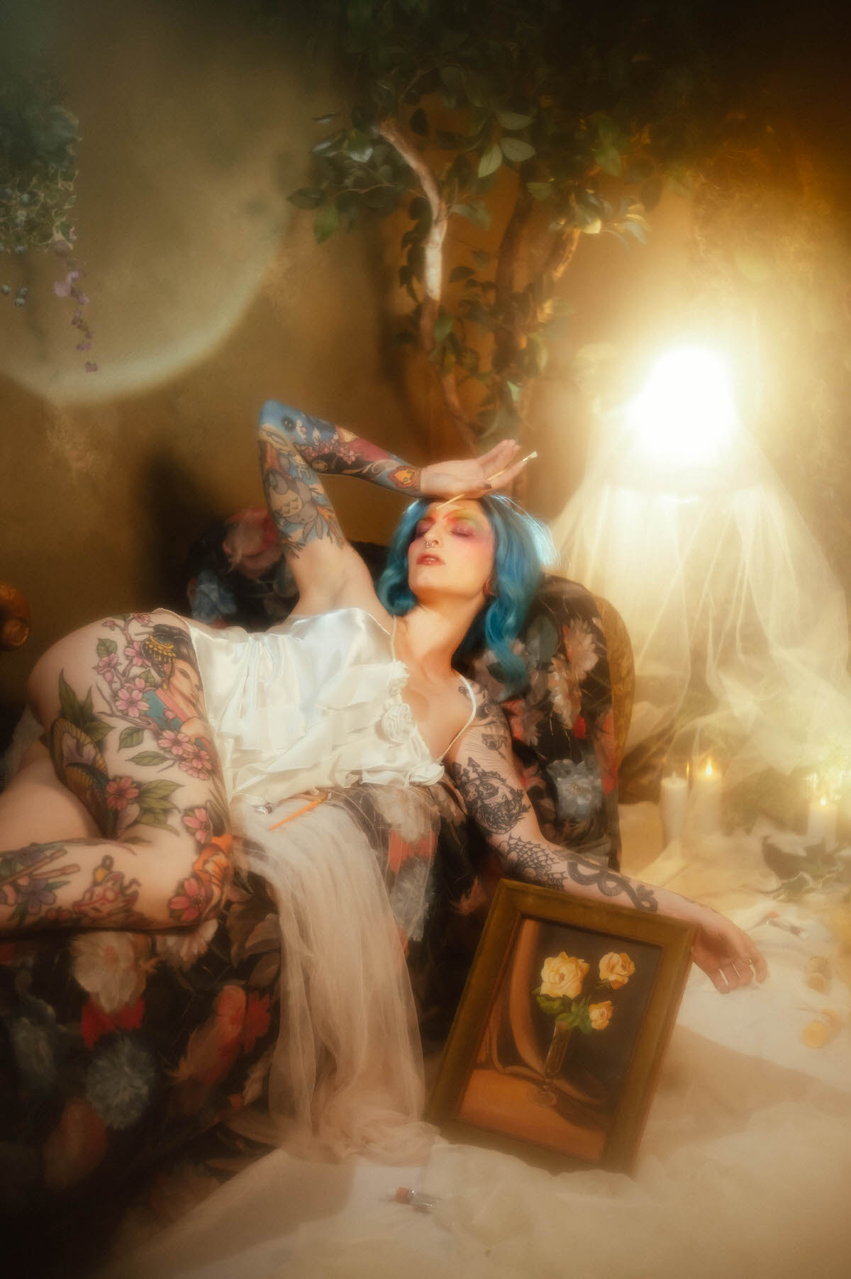 A woman with blue hair reclining on a floral chaise with one leg raised and eyes closed, an ivory slip dress falling open, and a gilt framed rose painting propped nearby during this artist boudoir photoshoot at Royal Lune Photo.