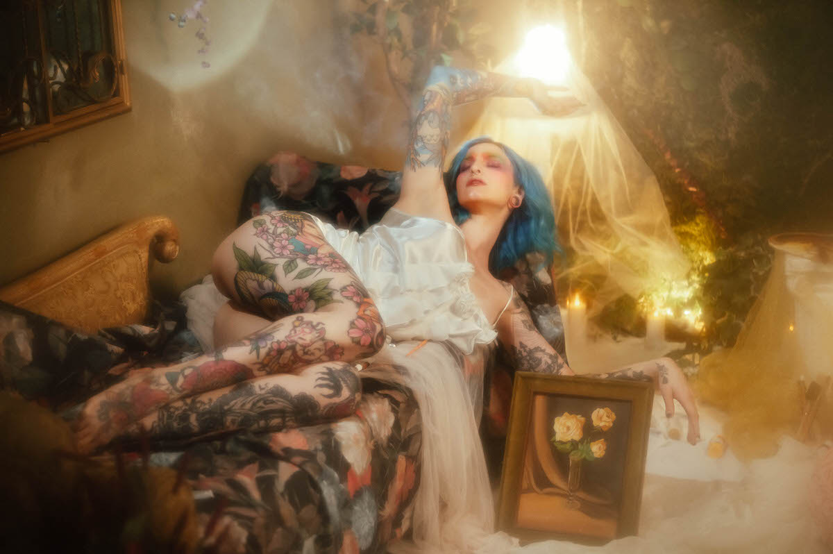 A woman with blue hair reclines on a floral upholstered chaise with one leg raised, wearing an ivory slip dress, beside a gilt frame holding a yellow rose painting during this artist boudoir photoshoot at Royal Lune Photo.