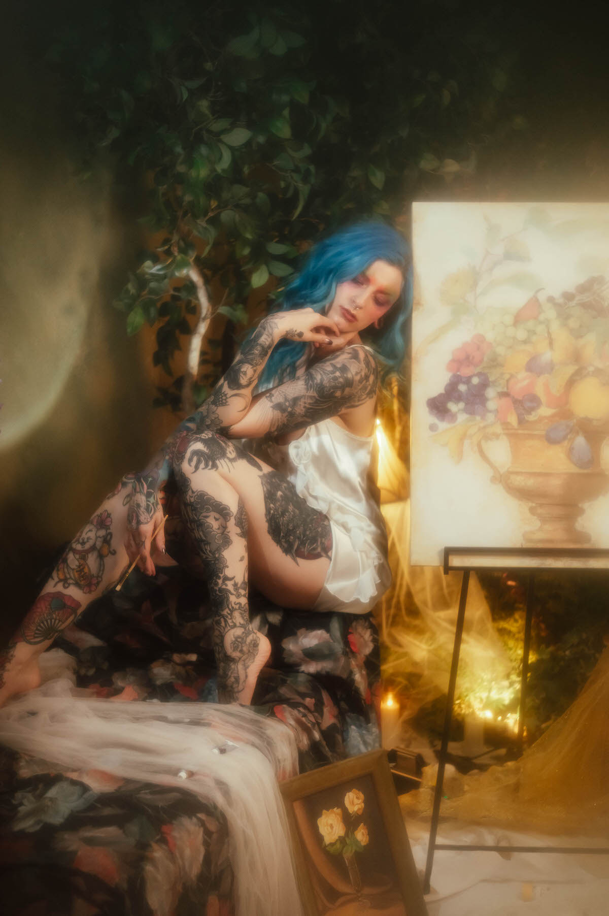 A woman in an ivory slip dress kneels on a floral chaise holding a paintbrush while facing a fruit and urn still life canvas on an easel, photographed by a Dallas fine art photographer at Royal Lune Photo.