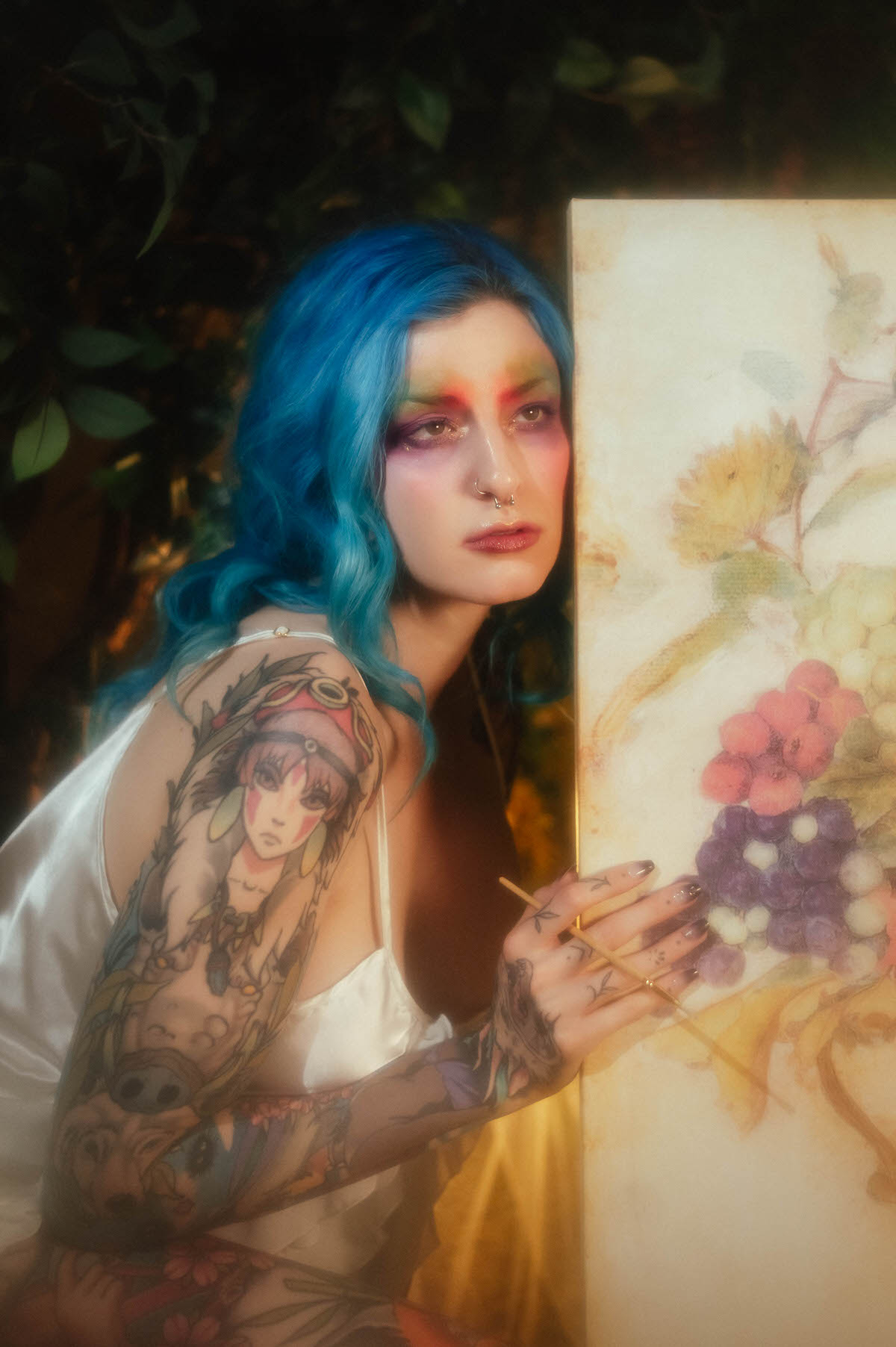A woman with blue hair rests her head against a fruit and floral still life canvas while touching it with a paintbrush, her tattooed shoulder visible beneath an ivory slip dress during this creative portrait photography session at Royal Lune Photo.