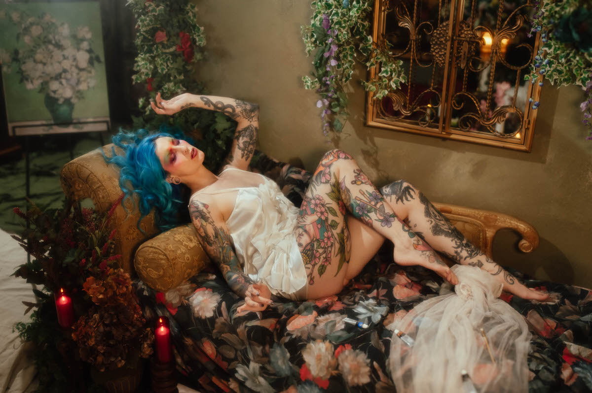 A woman with blue hair reclines on a gold upholstered chaise in an ivory slip dress beneath an ornate gold mirror, with a floral still life canvas and lit red candles nearby during this artist boudoir photoshoot at Royal Lune Photo.