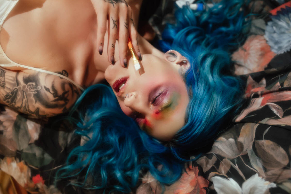 Close up of a woman with blue hair lying against floral fabric, holding a paintbrush near her lips with eyes closed, photographed by a Dallas fine art photographer at Royal Lune Photo.
