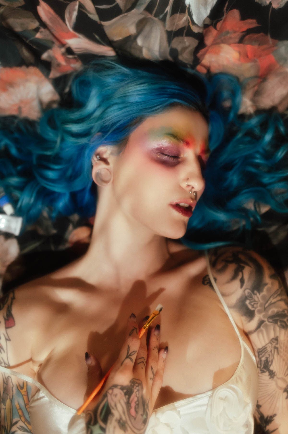 Overhead shot of a woman with blue hair fanned across a floral chaise, eyes closed, holding a paintbrush against her own chest during this fine art nude photography session at Royal Lune Photo.