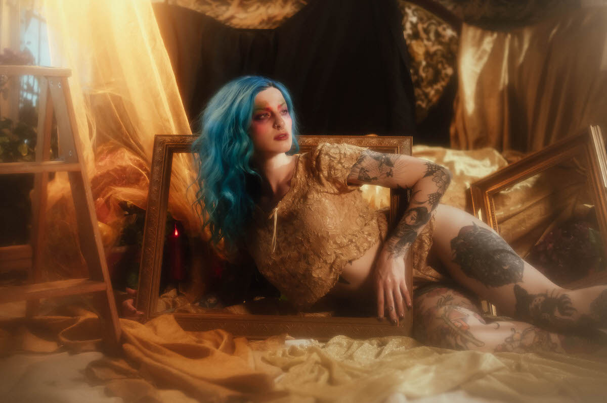 A woman with blue hair reclines with her torso framed inside a gold picture frame, a wooden step ladder and gold satin drapery in the background during this artist boudoir photoshoot at Royal Lune Photo.