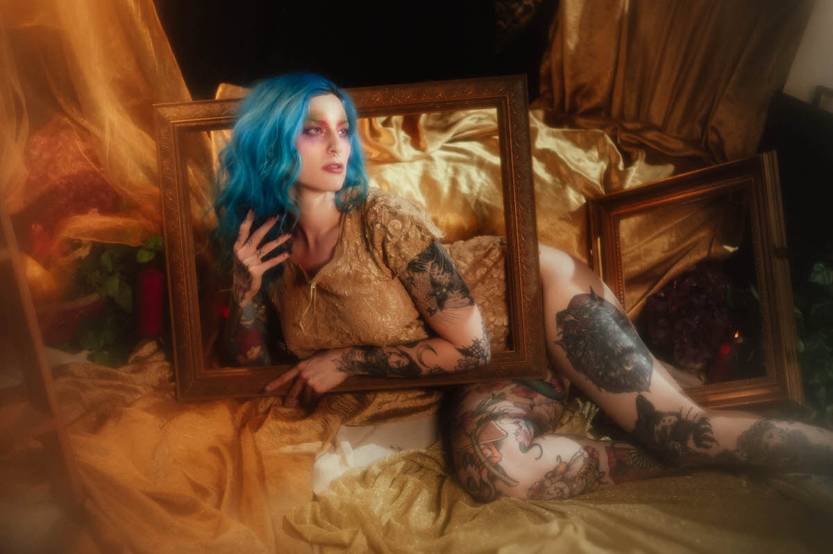 A woman with blue hair reclines against gold satin drapery with one hand raised to her hair, holding the edge of a gold picture frame during this artist boudoir photoshoot at Royal Lune Photo.