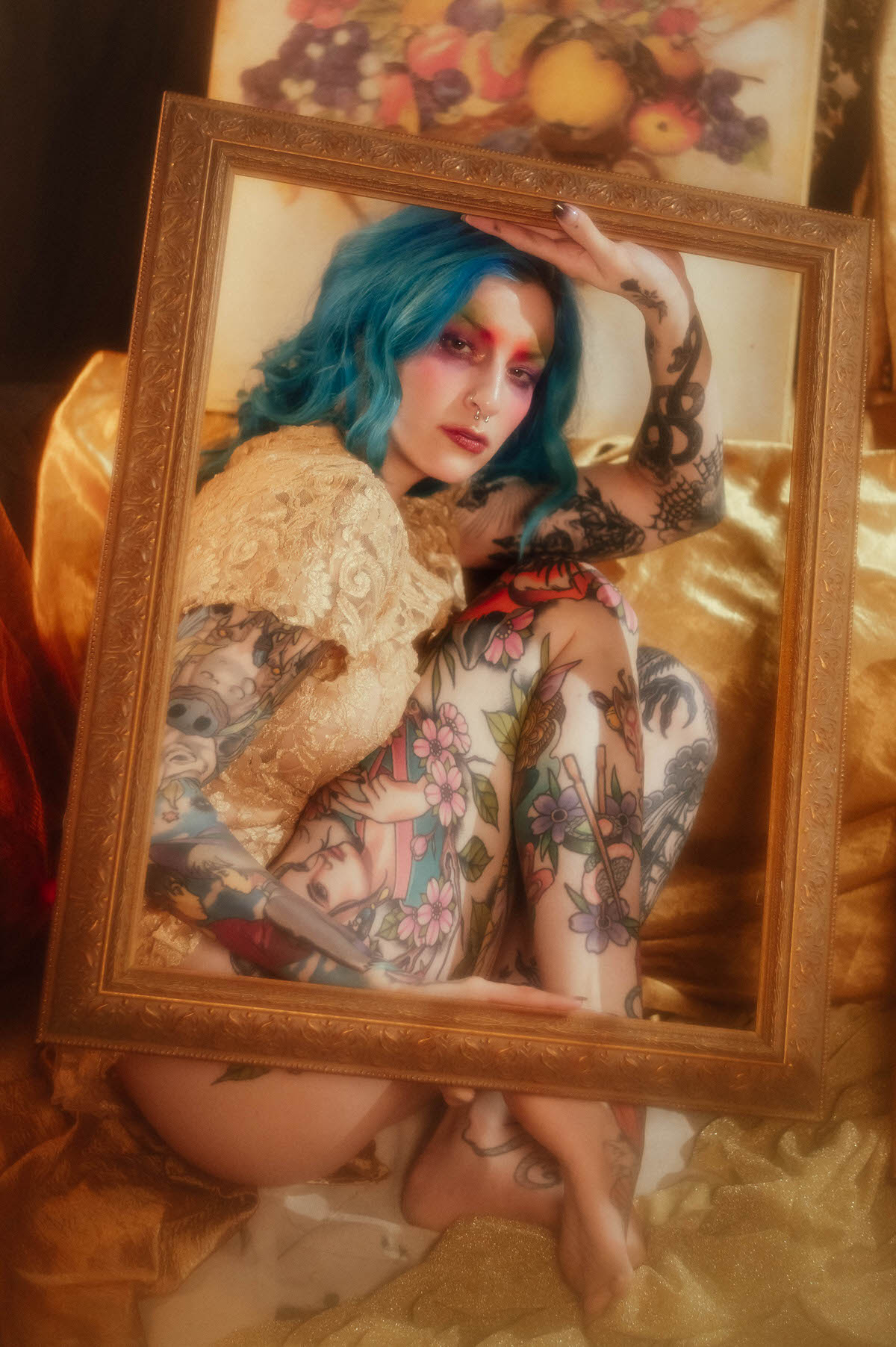 A woman in a gold lace top sits with her knees drawn up, full leg tattoos visible, viewed through a gold picture frame during this creative portrait photography session at Royal Lune Photo.