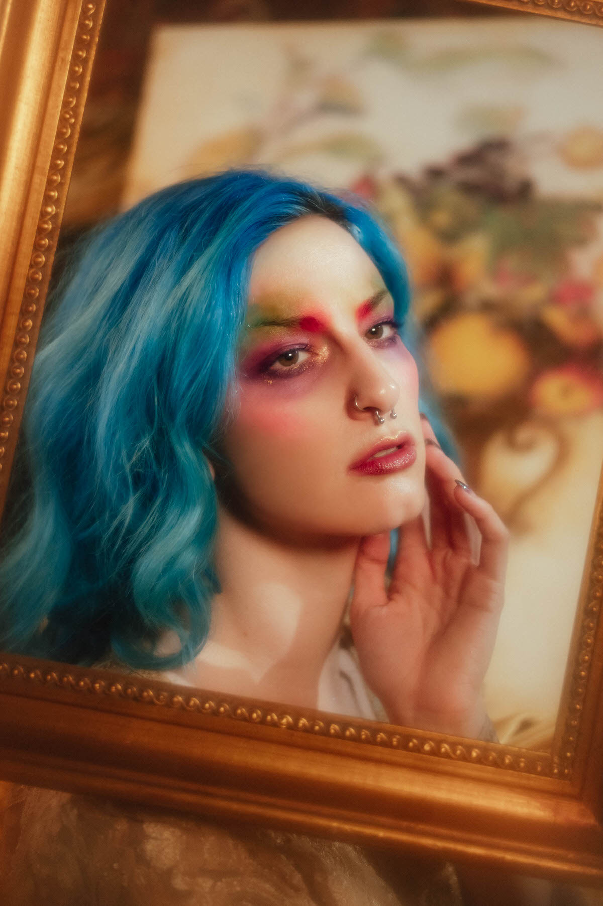Close up portrait of a woman with blue hair resting her hand near her chin, viewed through a gold picture frame with a blurred floral still life behind her during this fine art nude photography session at Royal Lune Photo.