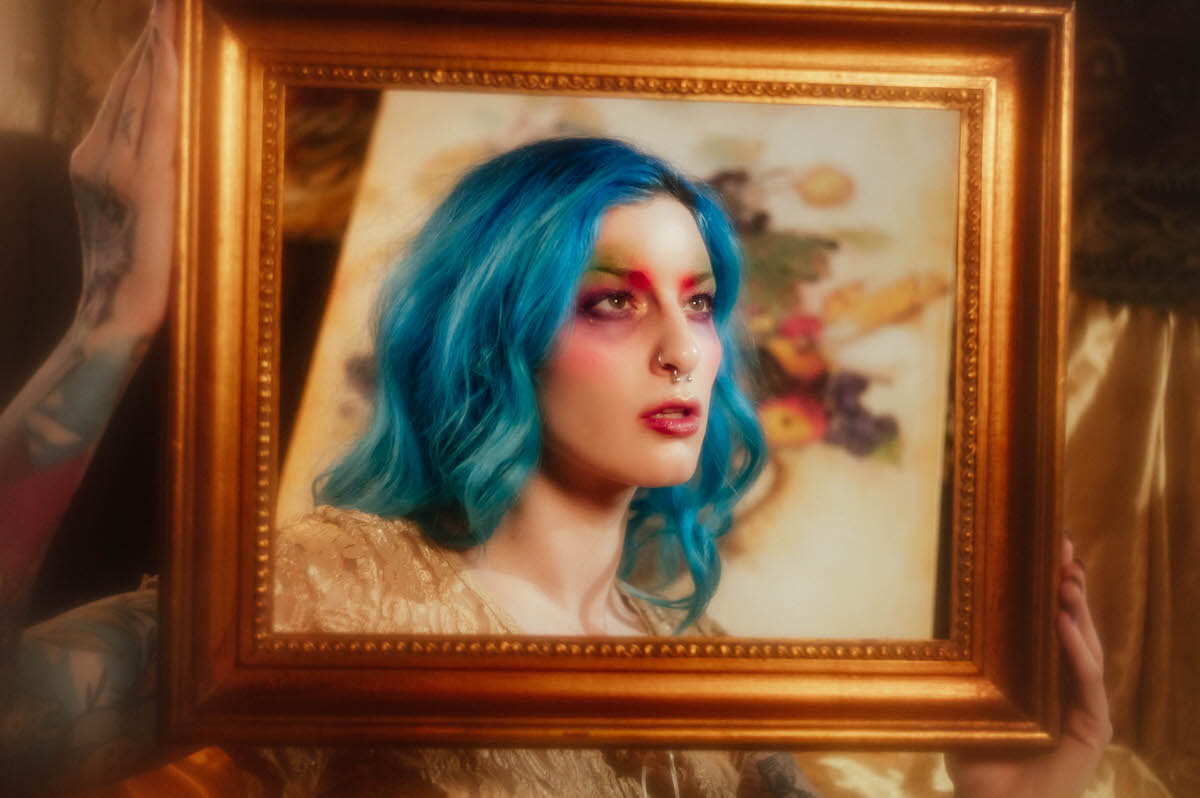 Side profile portrait of a woman with blue hair viewed through a gold picture frame, with a soft focus floral still life painting behind her during this artist boudoir photoshoot at Royal Lune Photo.