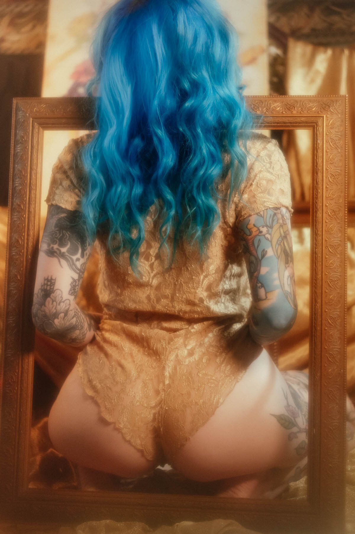 Rear view of a woman with blue hair kneeling in a gold lace two piece, viewed through a gold picture frame, tattooed arms visible on both sides during this artist boudoir photoshoot at Royal Lune Photo.
