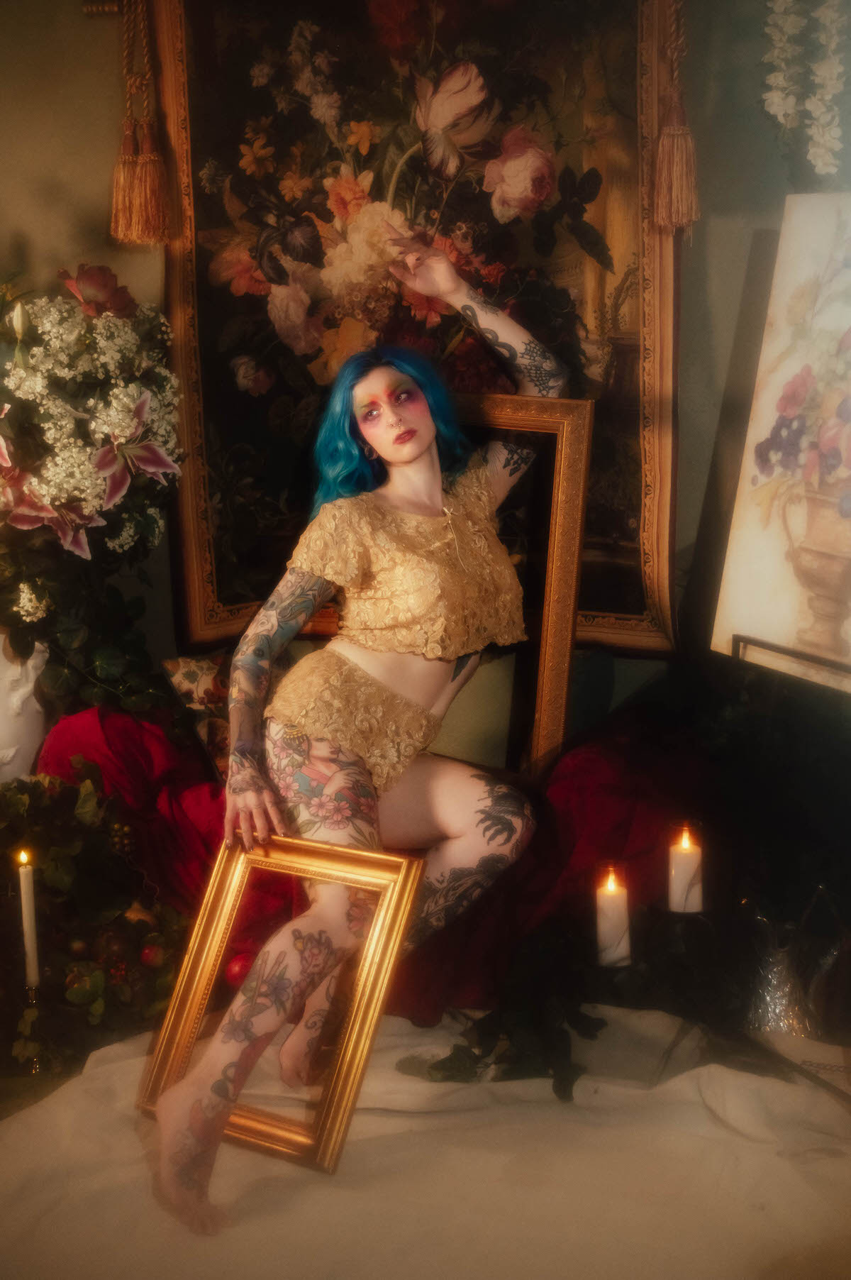 A woman in a gold lace two piece sits on an ornate chair holding a gold frame propped against her legs, flanked by lit candles and a floral tapestry backdrop during this artist boudoir photoshoot at Royal Lune Photo.