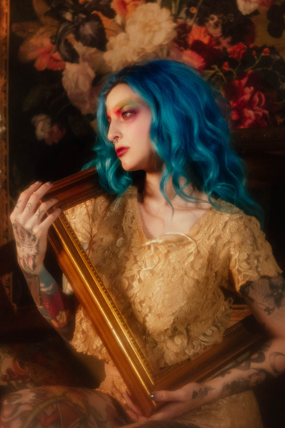 Profile portrait of a woman with blue hair holding a wooden picture frame against her shoulder, wearing a gold lace top, with a floral tapestry backdrop during this artist boudoir photoshoot at Royal Lune Photo.