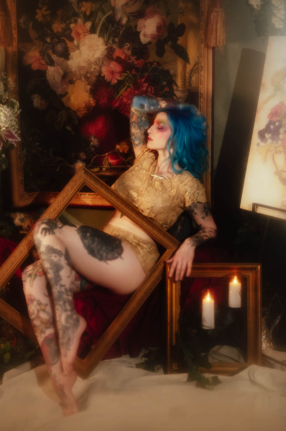 A woman with blue hair poses with her legs bent, viewed through a gold frame propped at an angle, with a floral tapestry and a smaller frame holding two candles nearby during this fine art nude photography session at Royal Lune Photo.