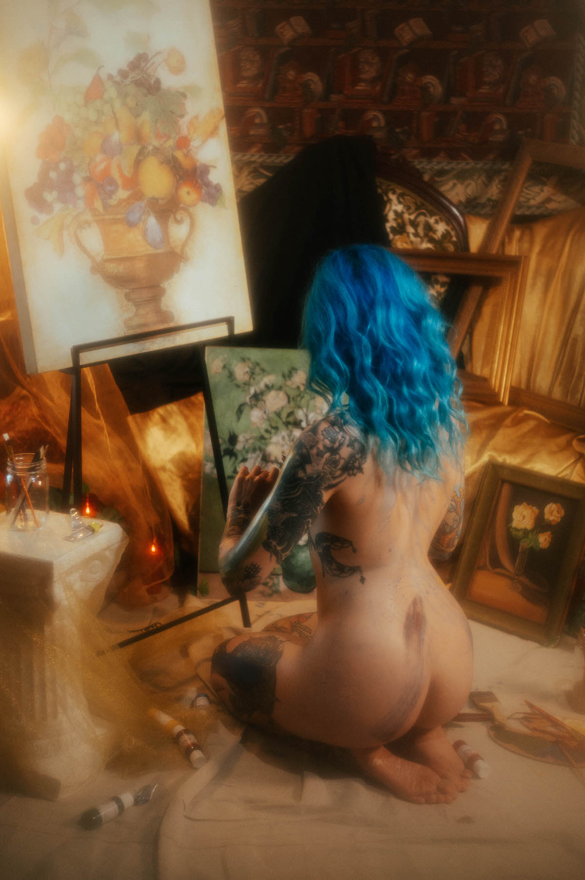 Rear view of a nude woman with blue hair kneeling before a fruit and urn still life canvas, hand raised toward a green floral canvas beside it, during this fine art nude photography session at Royal Lune Photo.