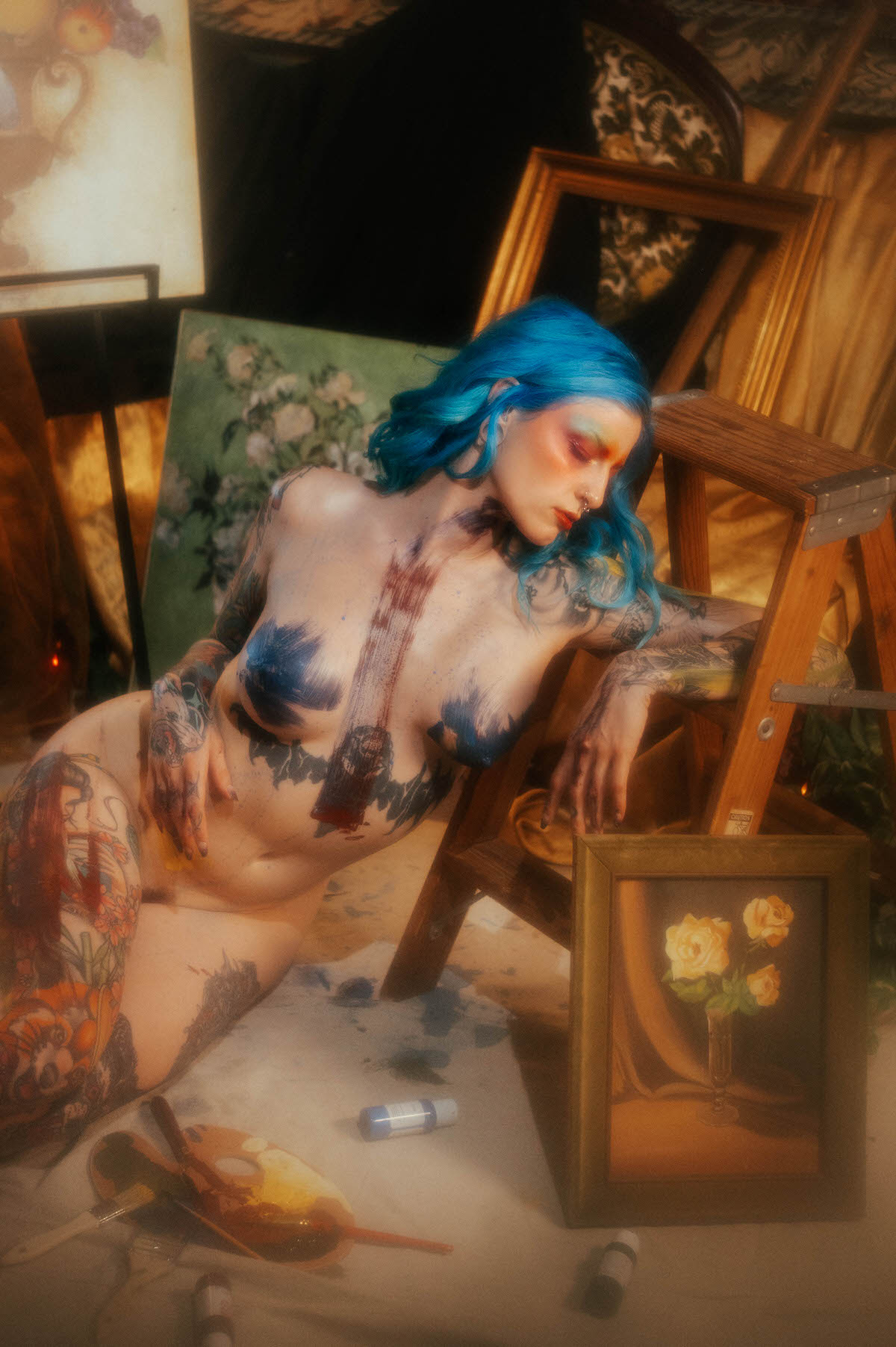 A nude woman with blue hair leans against a wooden step ladder, blue paint and a dark stripe across her chest and torso, with a green floral canvas behind her during this artist boudoir photoshoot at Royal Lune Photo.