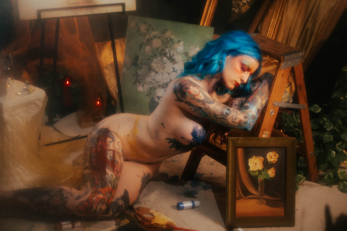A nude woman with blue hair curls into a wooden step ladder, tattooed arms crossed near her face, a smudge of yellow paint on her hip, photographed by a Dallas fine art photographer at Royal Lune Photo.