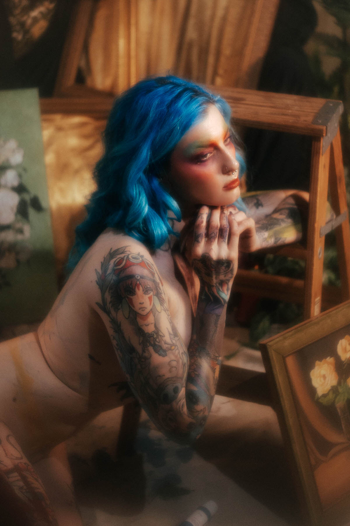 Close up of a woman with blue hair resting her chin on a tattooed hand near a wooden step ladder, a gold framed rose painting visible nearby during this body paint photoshoot at Royal Lune Photo.