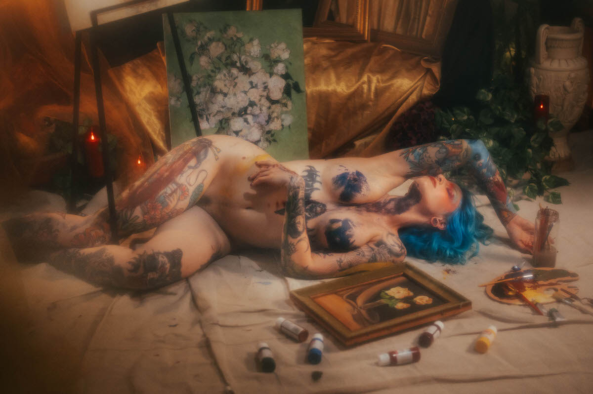 A nude woman with blue hair reclines with knees bent, blue paint brushed across her chest, surrounded by paint tubes, a palette, and a framed rose painting during this body paint photoshoot at Royal Lune Photo.