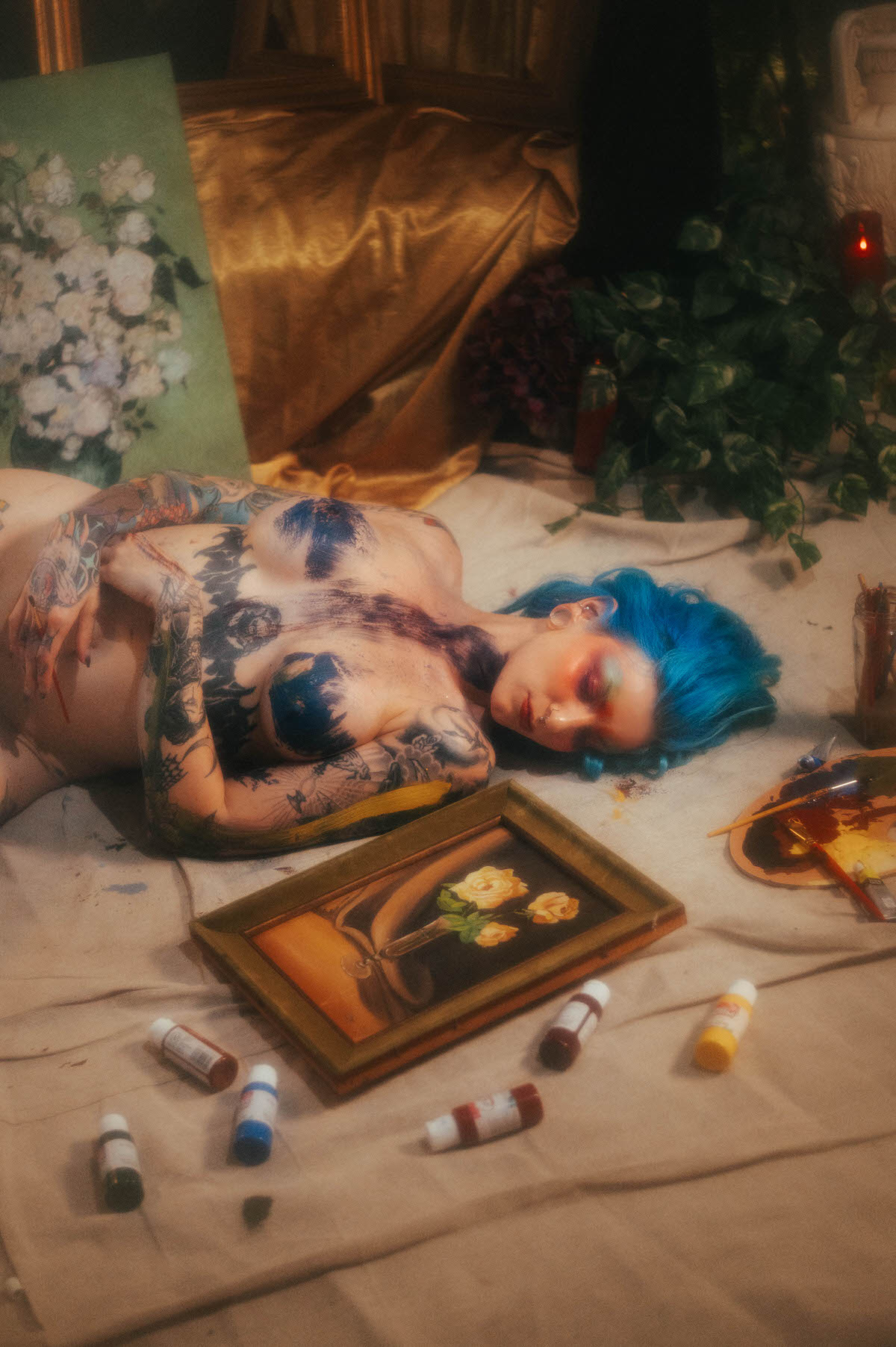 A nude woman with blue hair lies on a canvas drop cloth with blue and dark paint brushed across her chest, a framed rose still life and scattered paint tubes at her feet during this artist boudoir photoshoot at Royal Lune Photo.