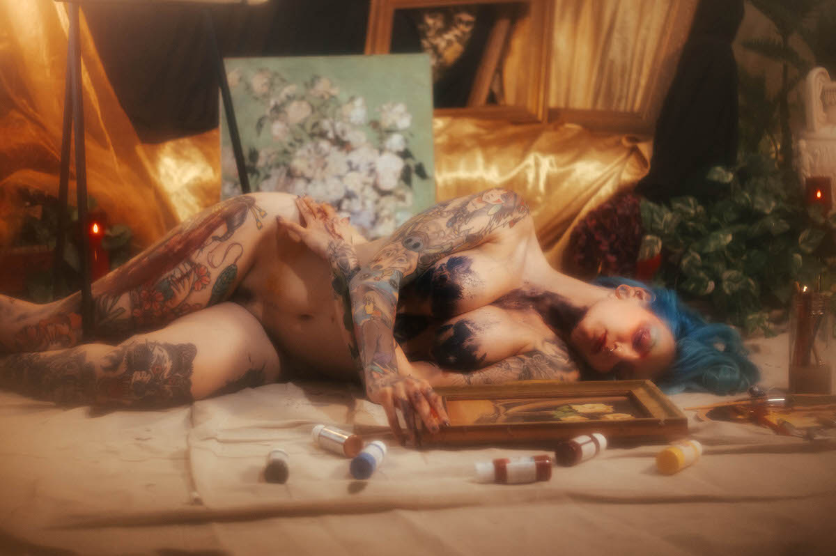 A nude woman with blue hair reclines on a canvas drop cloth, blue paint across her chest, reaching for a framed still life painting among scattered paint tubes during this artist boudoir photoshoot at Royal Lune Photo.