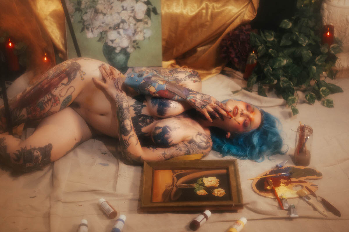 A nude woman with blue hair lies on her back with a paintbrush held to her lips, blue paint covering her chest, paint tubes scattered around her during this fine art nude photography session at Royal Lune Photo.