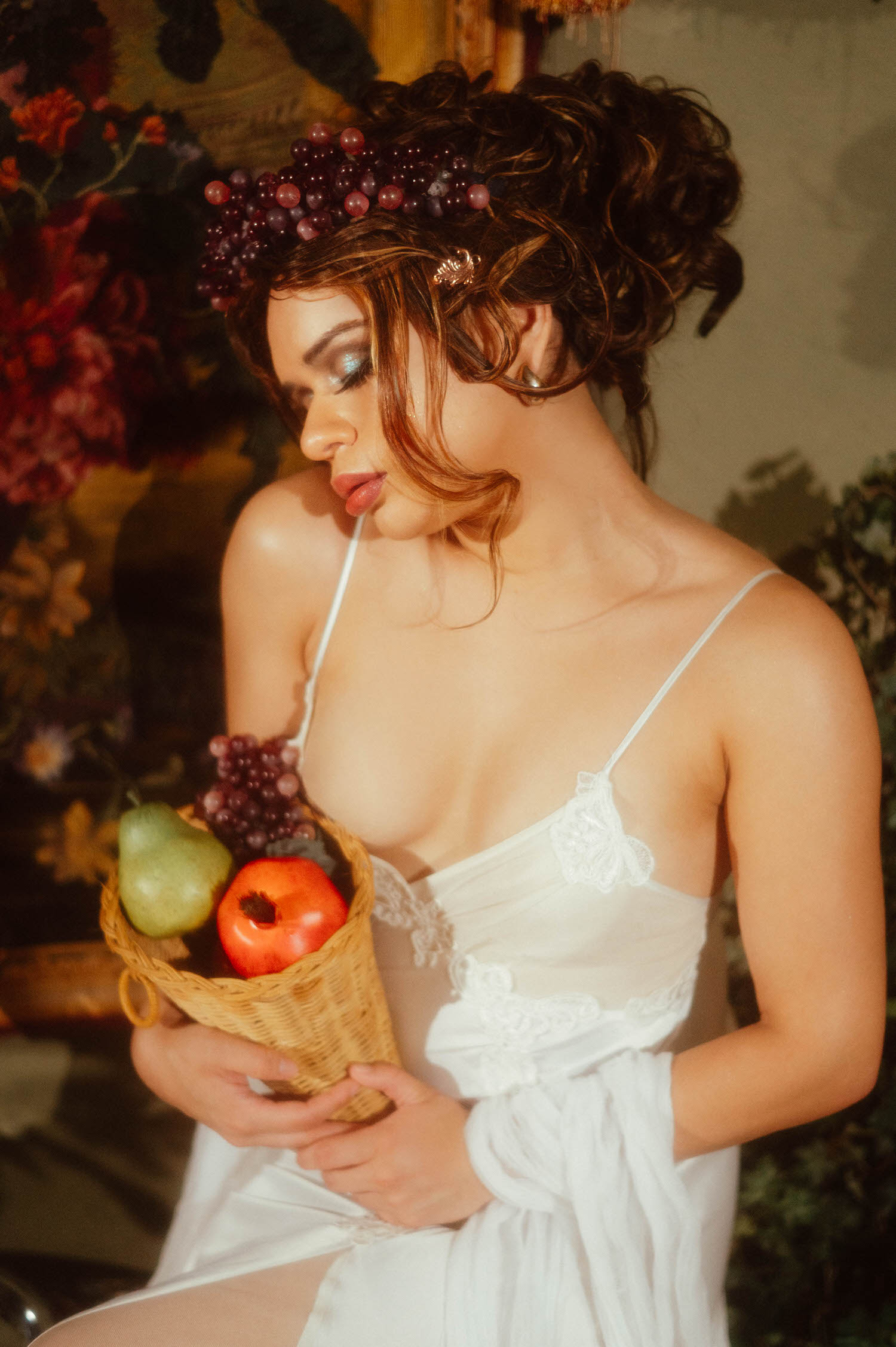 Profile portrait of a woman with a grape crown wig in this fine art photo studio setting, glancing downward toward a basket of pears and grapes, Royal Lune Photo.