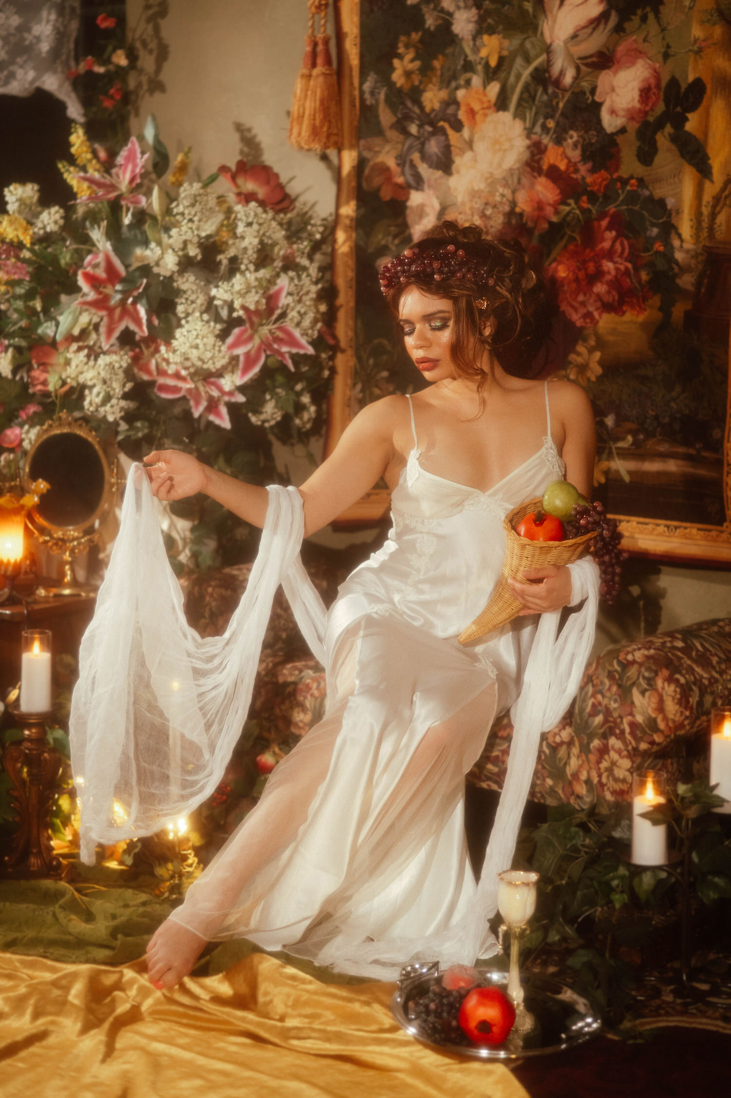 Woman in a flowing white gown extending sheer fabric outward during this mucha inspired photoshoot, seated among candles and floral tapestries, Royal Lune Photo.