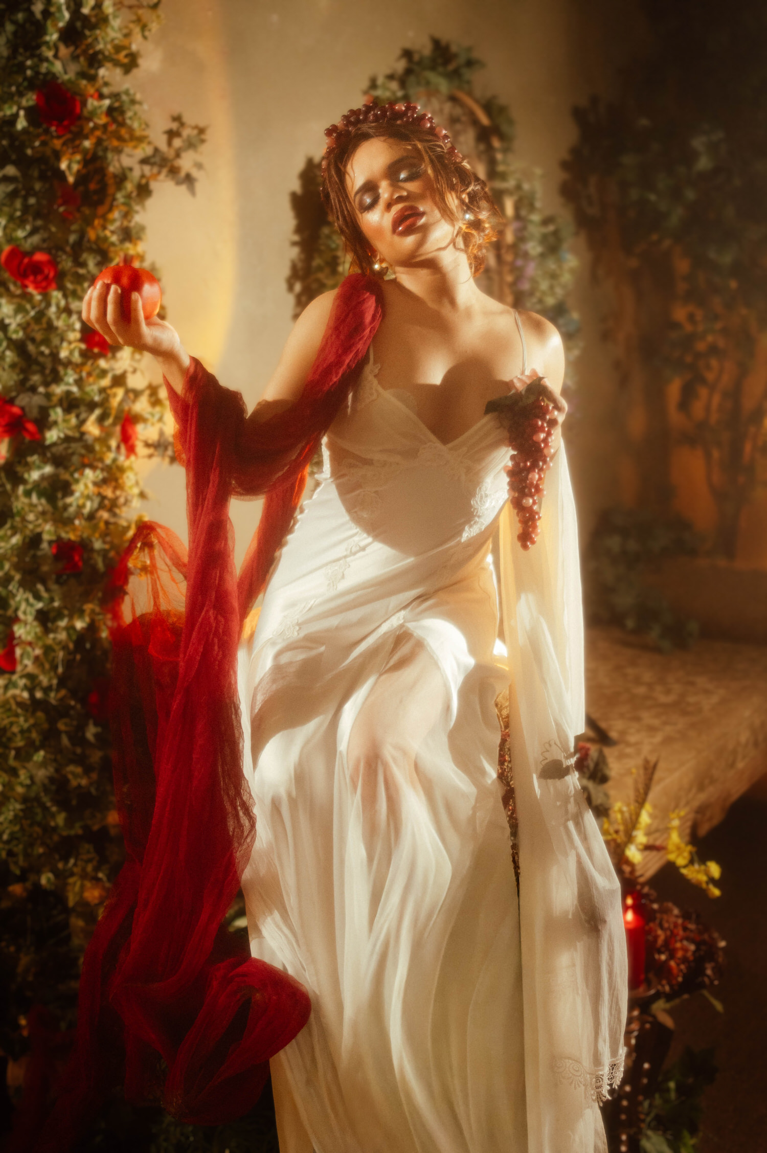 Woman in a vintage nightgown with red drapery cascading from her shoulder, holding a pomegranate and grapes against a rose covered backdrop, Royal Lune Photo.