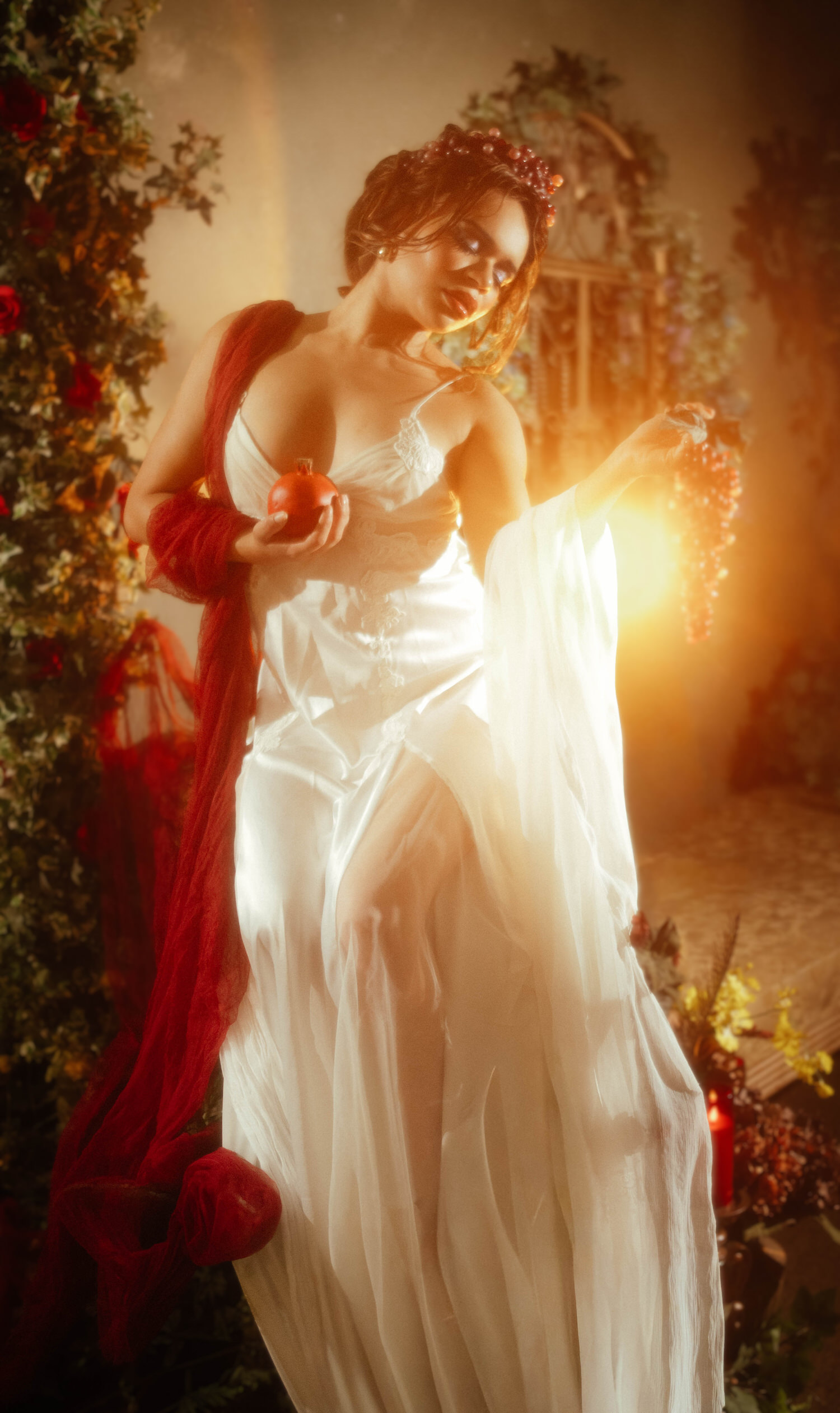 Woman with a grape crown and art nouveau styled wig holding a pomegranate and grapes in golden backlight during this art nouveau photography session, red drapery flowing from her shoulder, Royal Lune Photo.