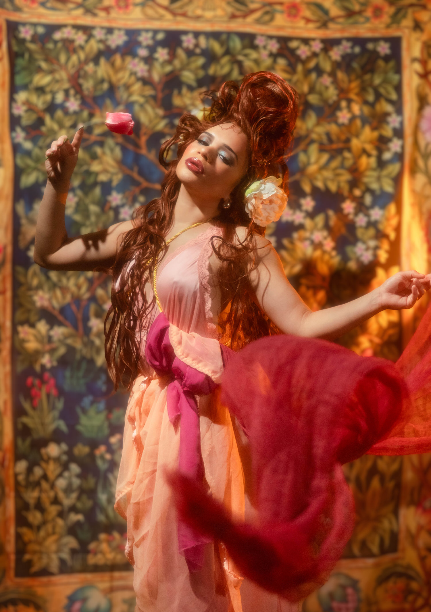 Woman holding a pink tulip into the air with red drapery swirling around her against a gold bordered tapestry backdrop, Royal Lune Photo.
