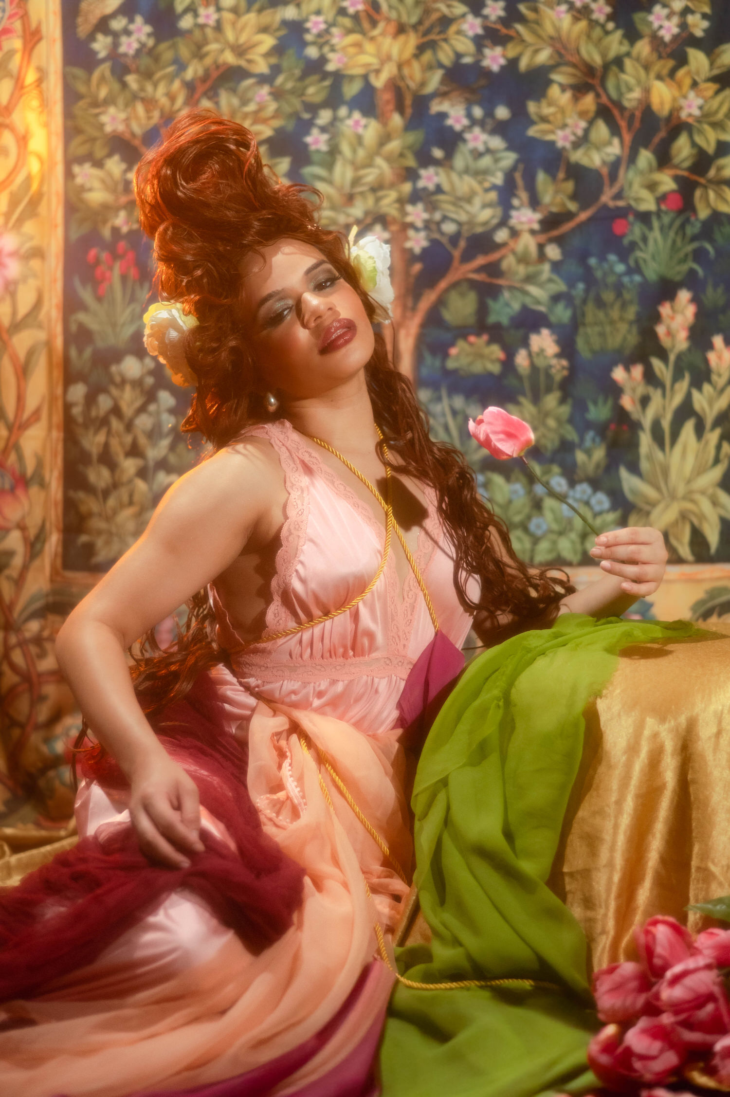 Woman seated on a chaise draped in green and gold fabric while holding a single pink tulip in this fine art photo studio setting, Royal Lune Photo.