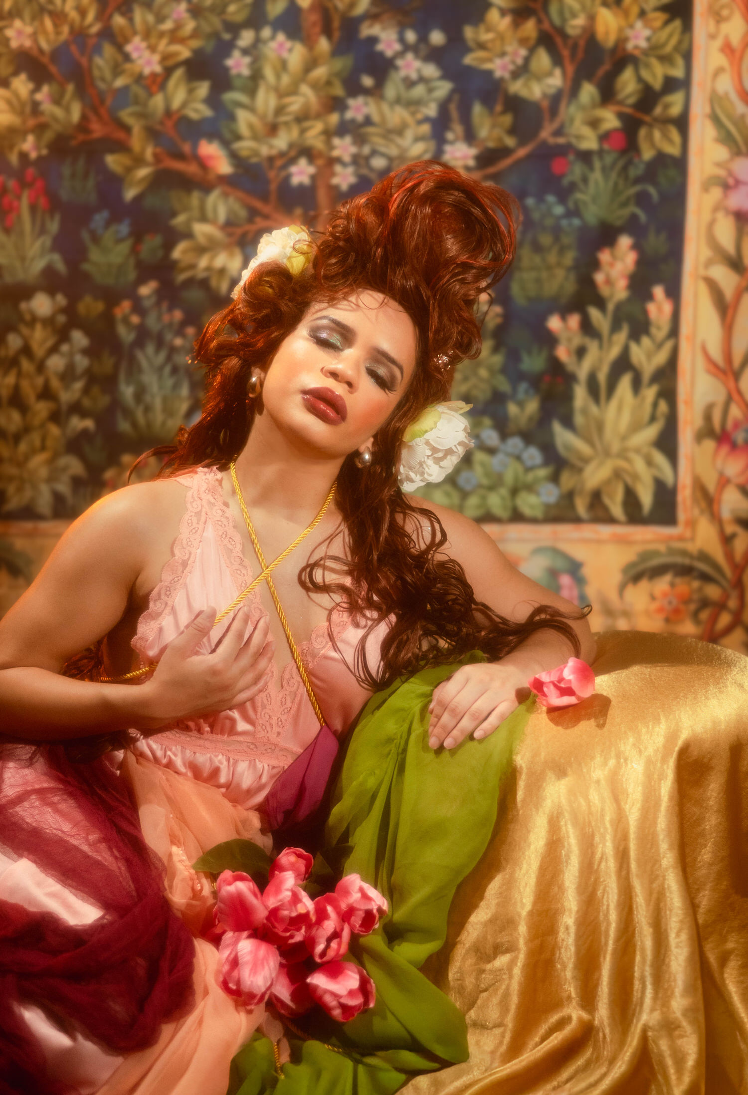 Woman resting against a golden chaise surrounded by pink roses and green drapery with one hand at her chest, Royal Lune Photo.