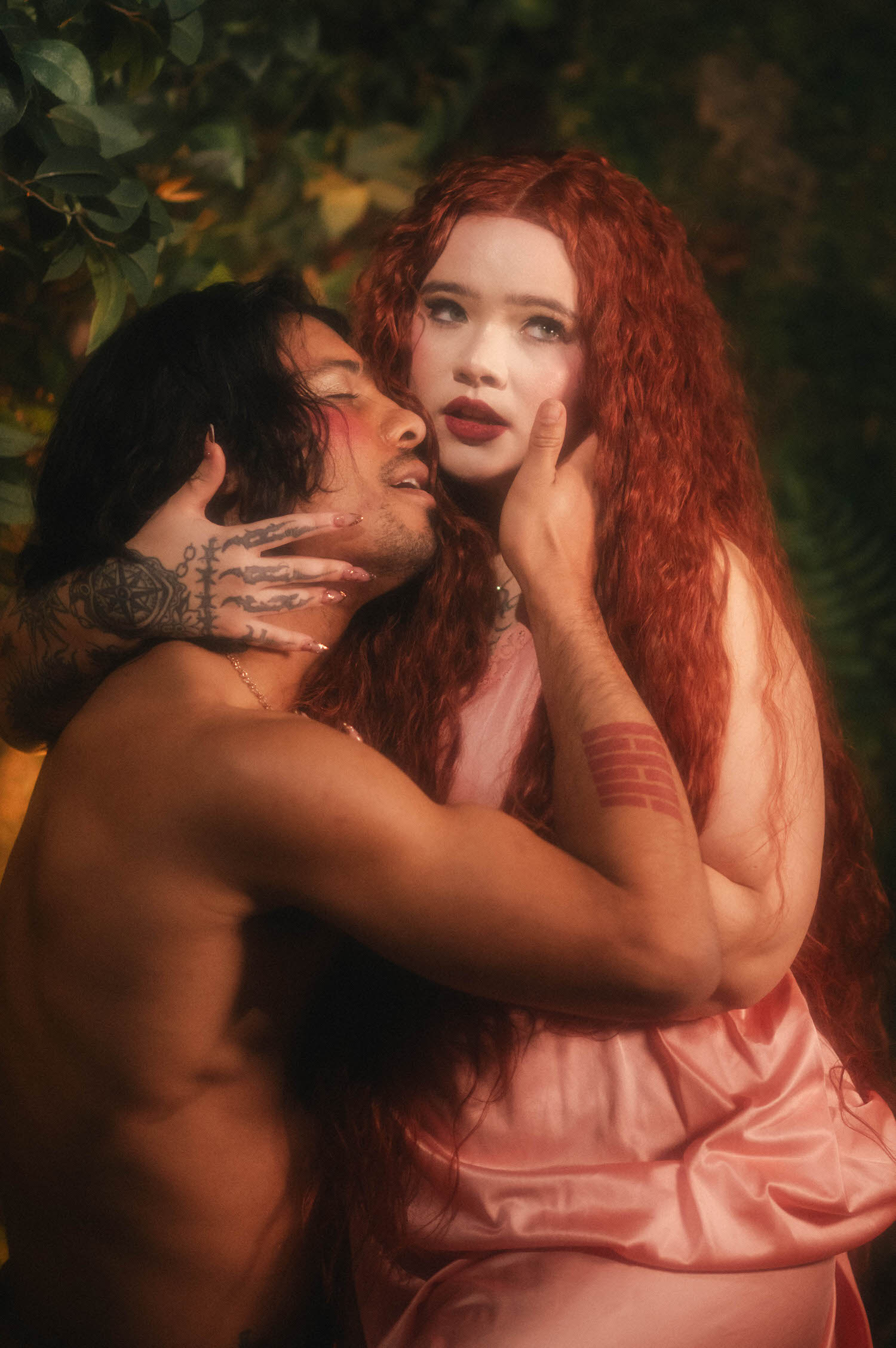 A statue inspired photoshoot pose captures a couple face to face in dappled greenery, the woman's hand resting against her partner's cheek as tattooed forearms frame the embrace. Royal Lune Photo