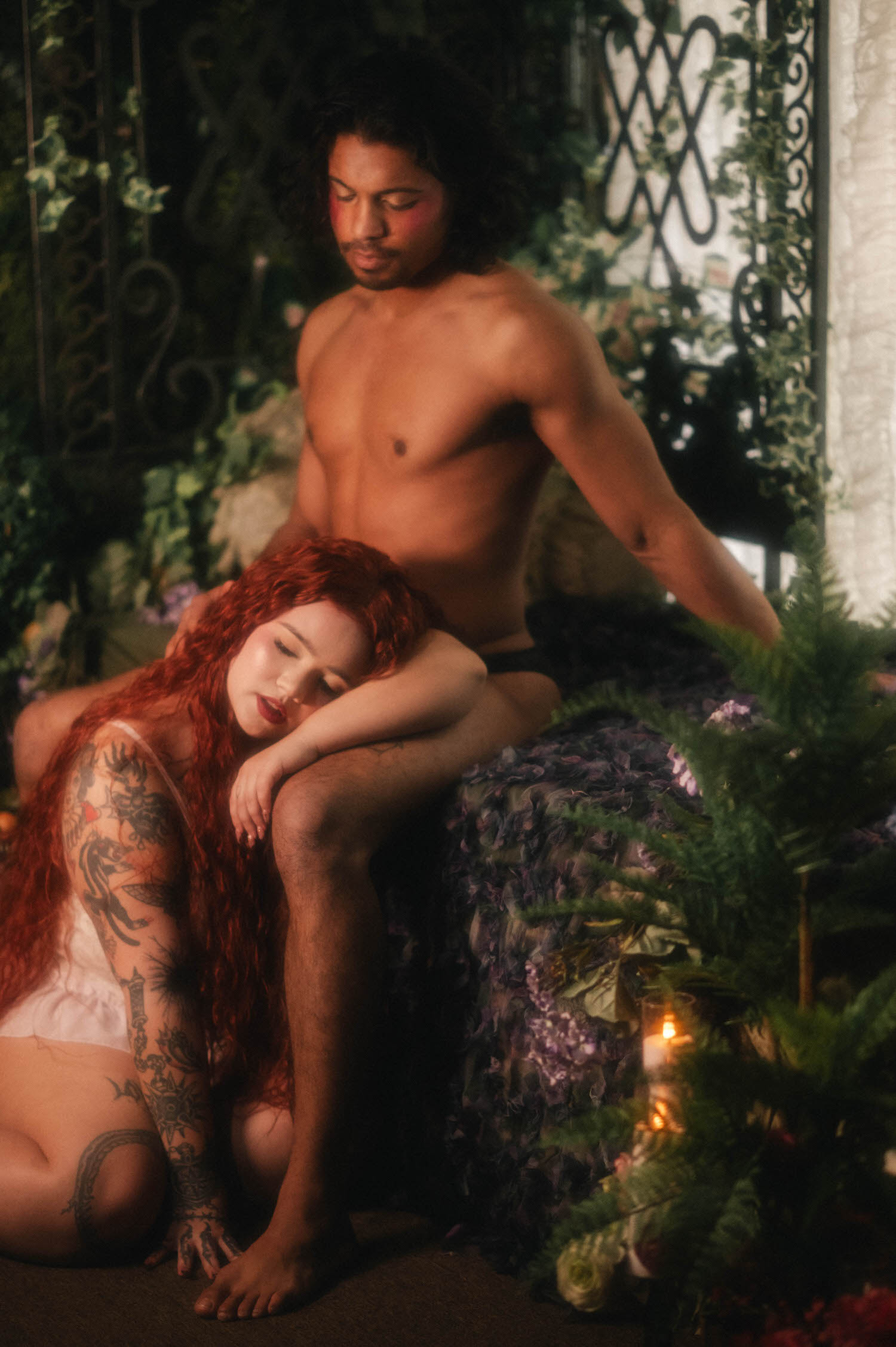 A Renaissance boudoir photoshoot moment shows a woman resting against her partner's leg, red curls pooling across his lap as candlelight glows beside wisteria blooms. Royal Lune Photo
