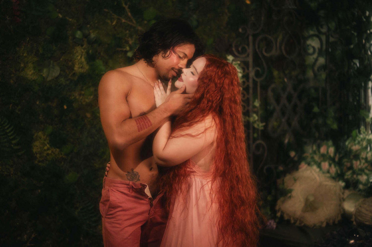 A Renaissance boudoir photoshoot featuring a couple embracing in front of a wrought iron gate, the woman's long auburn hair cascading over her shoulder as she leans into her partner's touch. Royal Lune Photo
