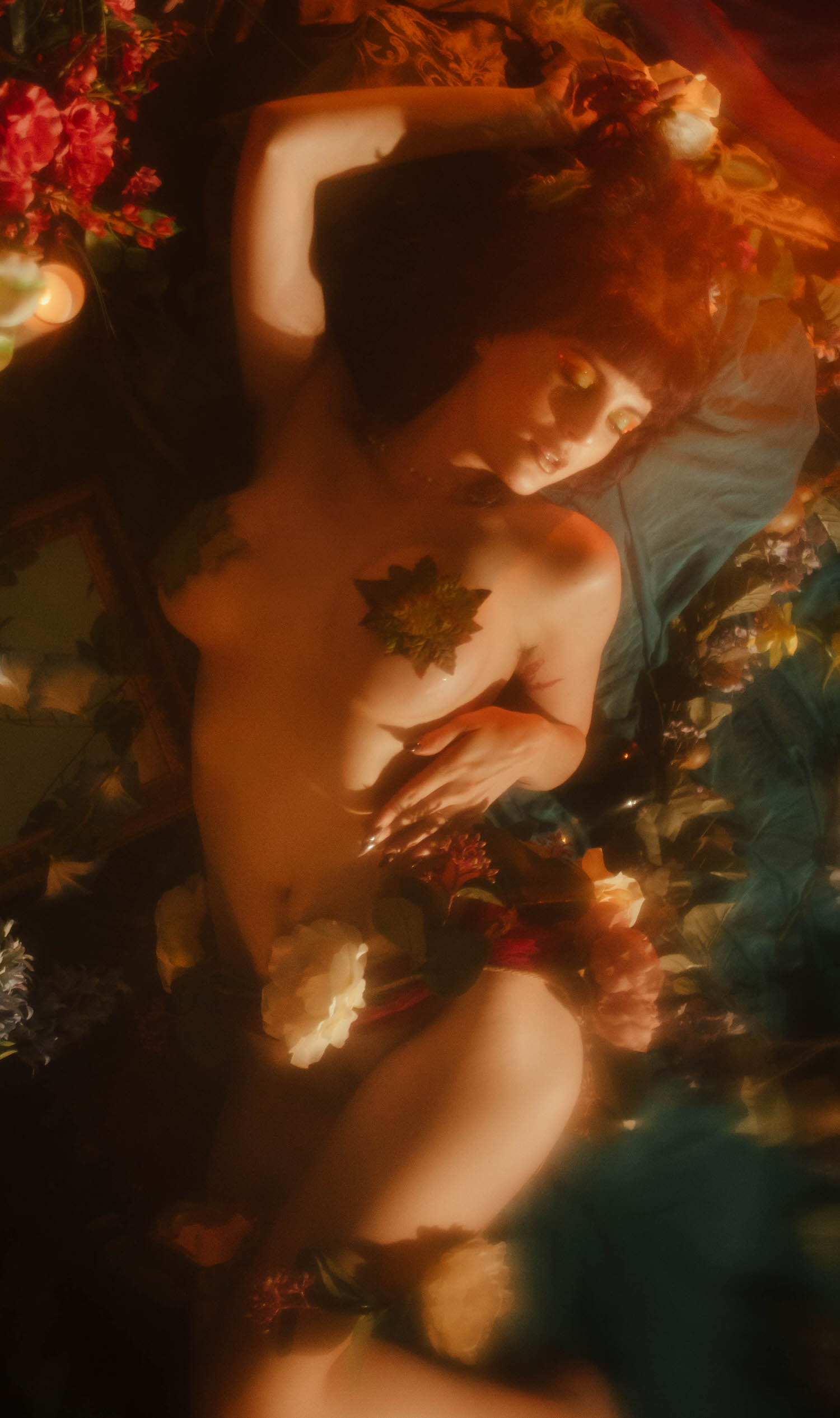 A model lies among red and cream roses on teal fabric, styled in the nude look with a pressed leaf detail, fog machine haze glowing warm around her, part of a whimsigoth aesthetic gallery at Royal Lune Photo.