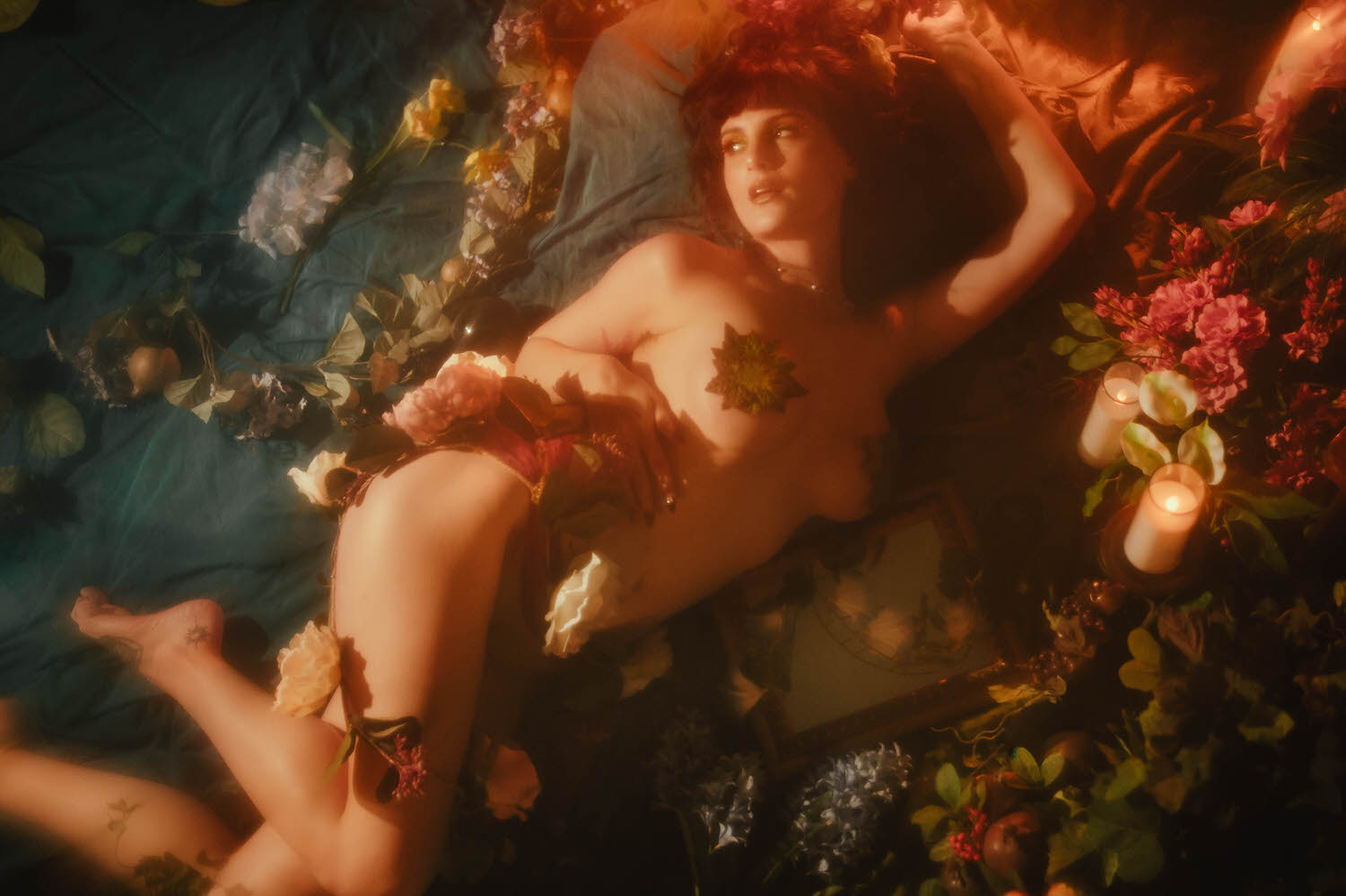 A model is laying down on teal fabric surrounded by pink and white florals and lit candles, styled in the nude look with pressed leaves, soft fog catching the warm light in a moody boudoir photoshoot at Royal Lune Photo.