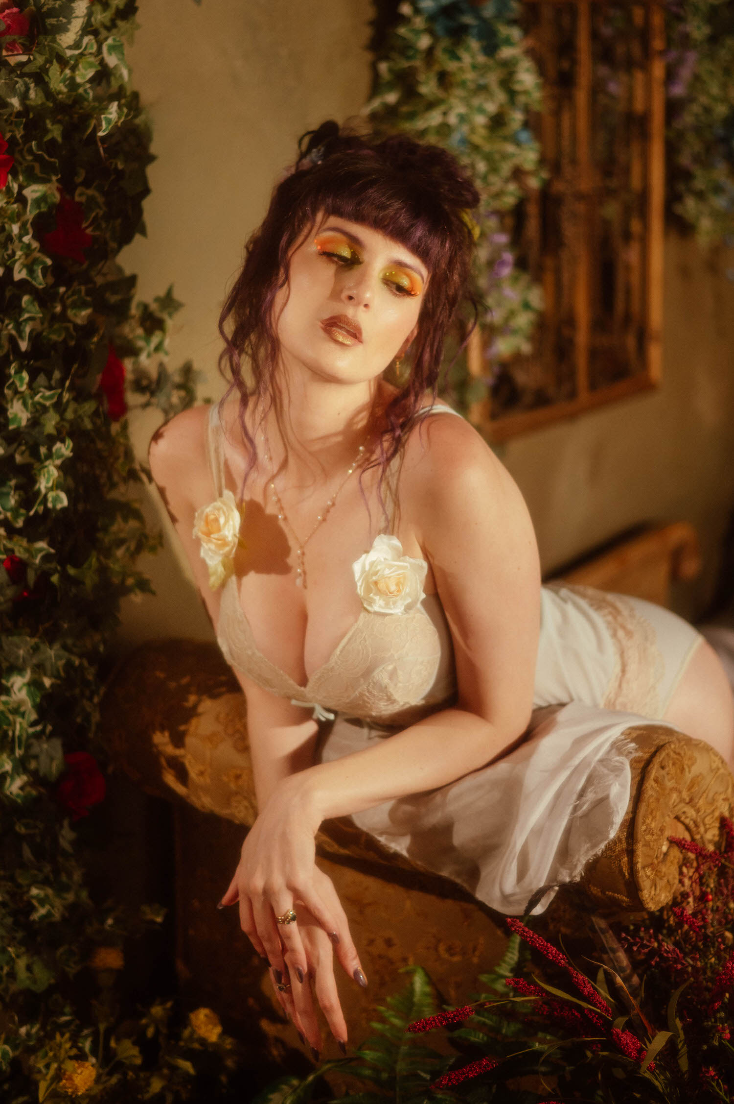 A model sits on an antique bench in a cream lace bodysuit with rose shoulder details, surrounded by ivy and red roses under warm golden light, styled at a Dallas Fort Worth photo studio for Royal Lune Photo.