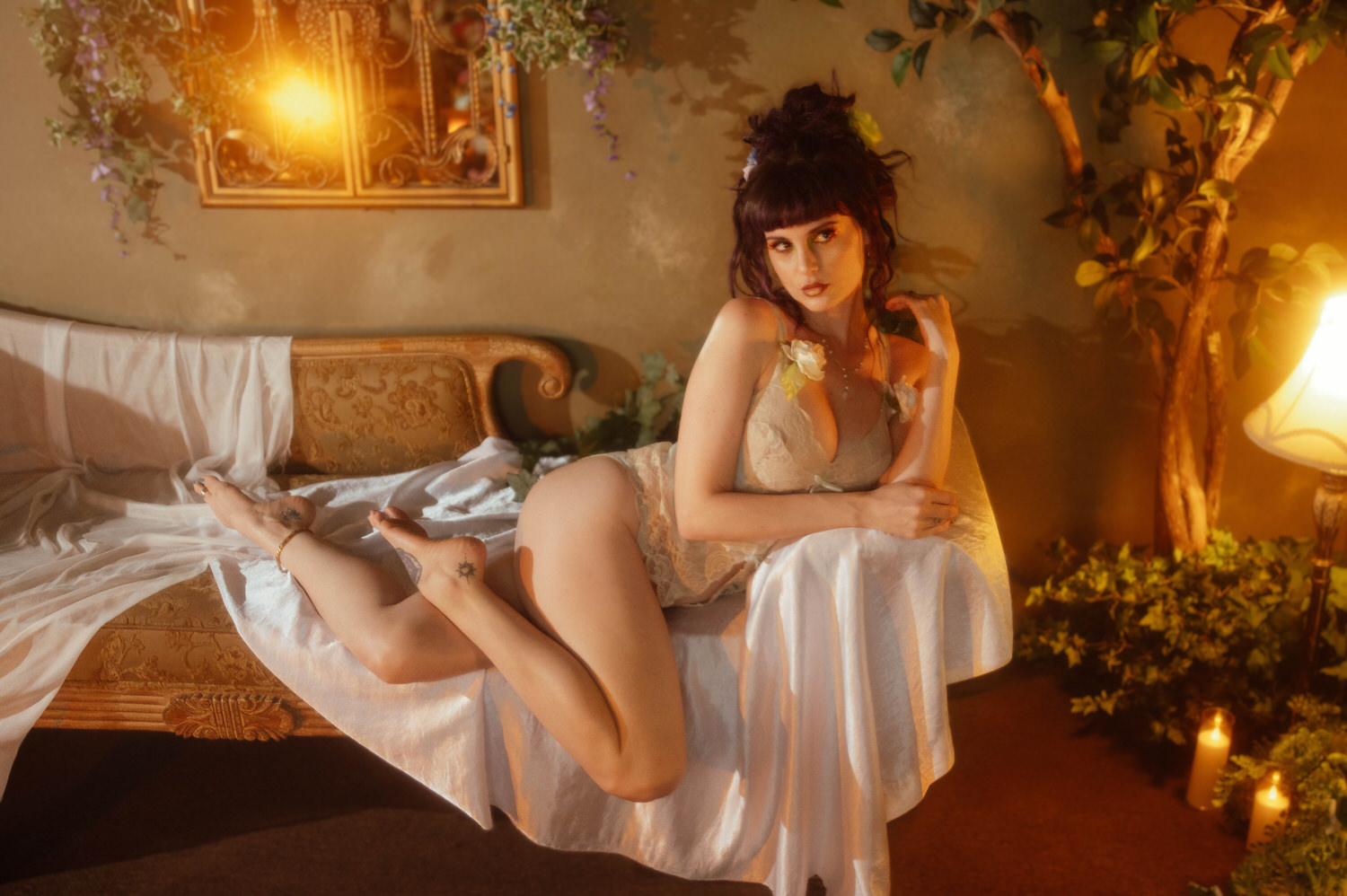 A model reclines across a white sheet with legs extended, wearing a cream lace bodysuit and looking over her shoulder near a lit candle and trailing ivy, styled by a Dallas Fort Worth boudoir photographer at Royal Lune Photo.