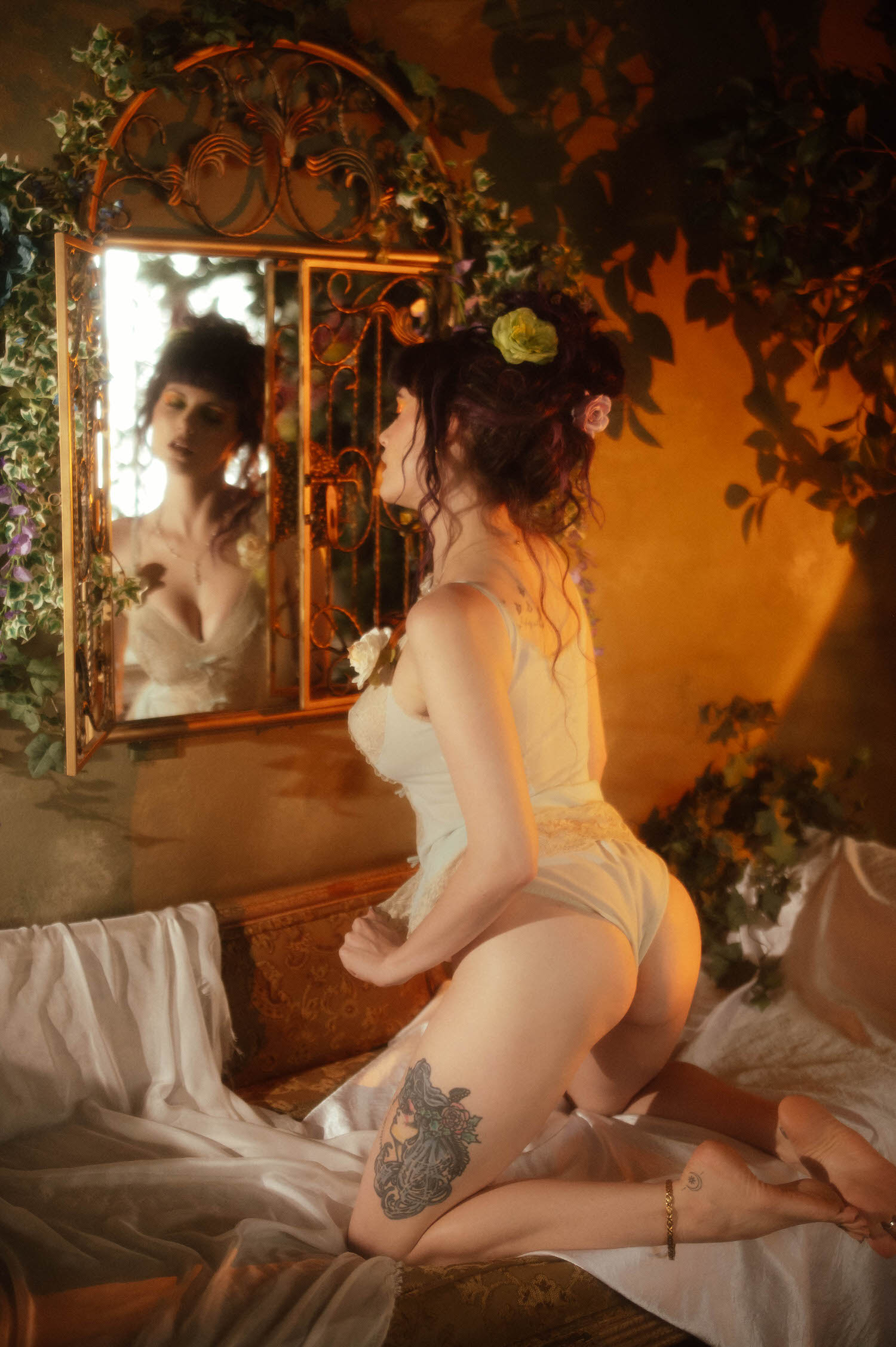 A model kneels facing an ornate gold mirror in a cream lace bodysuit, her reflection catching soft warm light against a wall lined with ivy, part of a moody boudoir photoshoot at Royal Lune Photo.