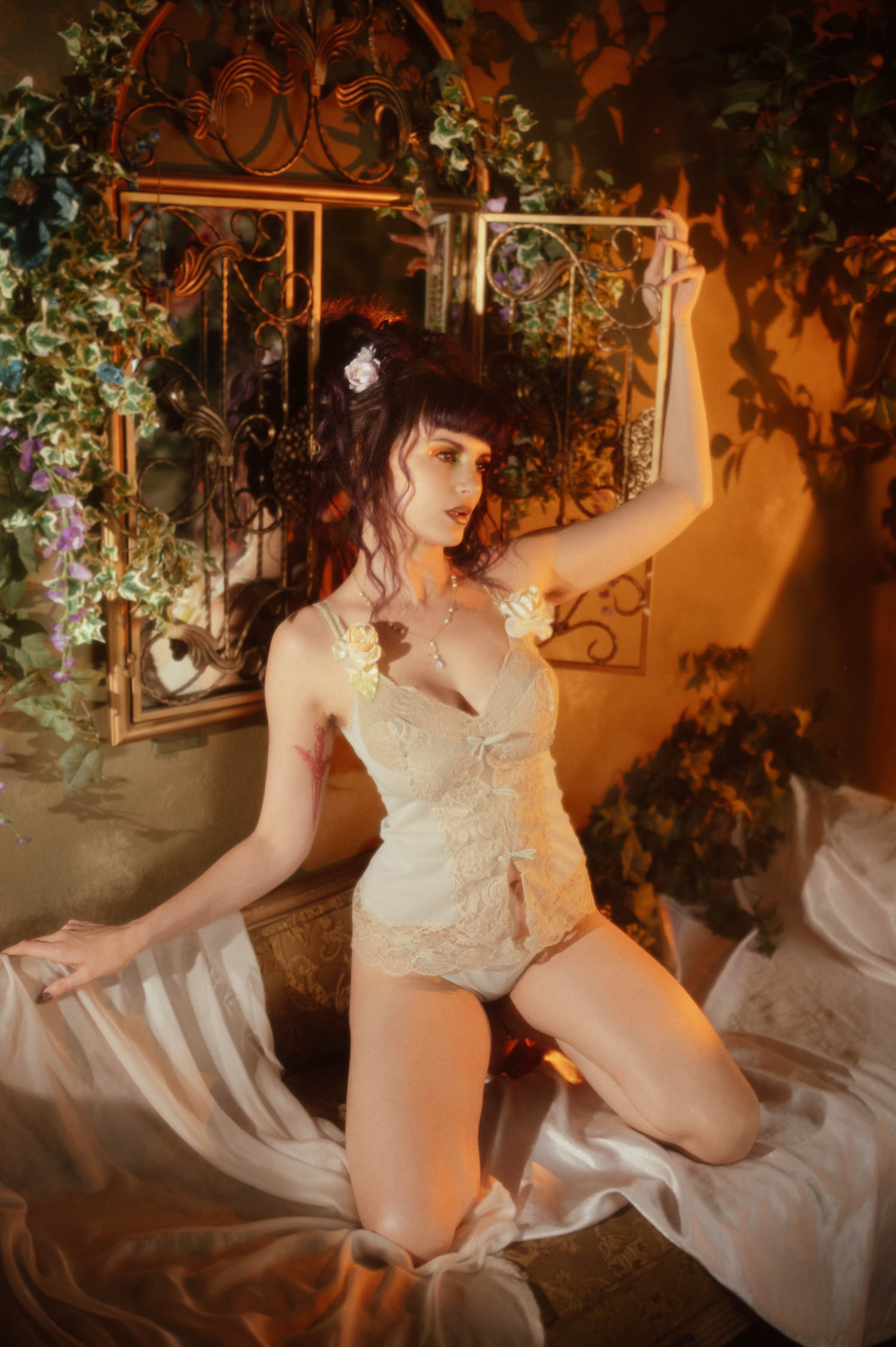 A model kneels on white sheer fabric in a vintage mint green lingerie set with rose shoulder straps, reaching toward an ornate mirror framed by ivy in warm golden light, part of a moody boudoir photoshoot at Royal Lune Photo.
