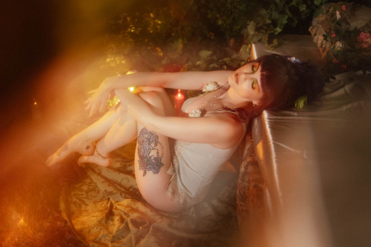 A model reclines on a gold brocade fabric in a cream lace bodysuit with rose strap details, her leg tattoo catching warm lens flare light, photographed during a moody boudoir photoshoot at Royal Lune Photo.
