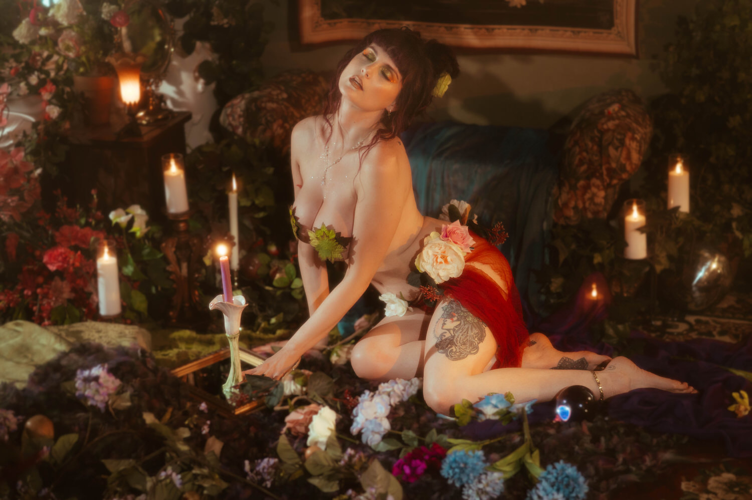 A model kneels among candles, roses, and hydrangeas in the nude look styled with pressed leaves and a sheer red fabric wrap, showing a detailed thigh tattoo in warm candlelight, part of a moody boudoir photoshoot at Royal Lune Photo.